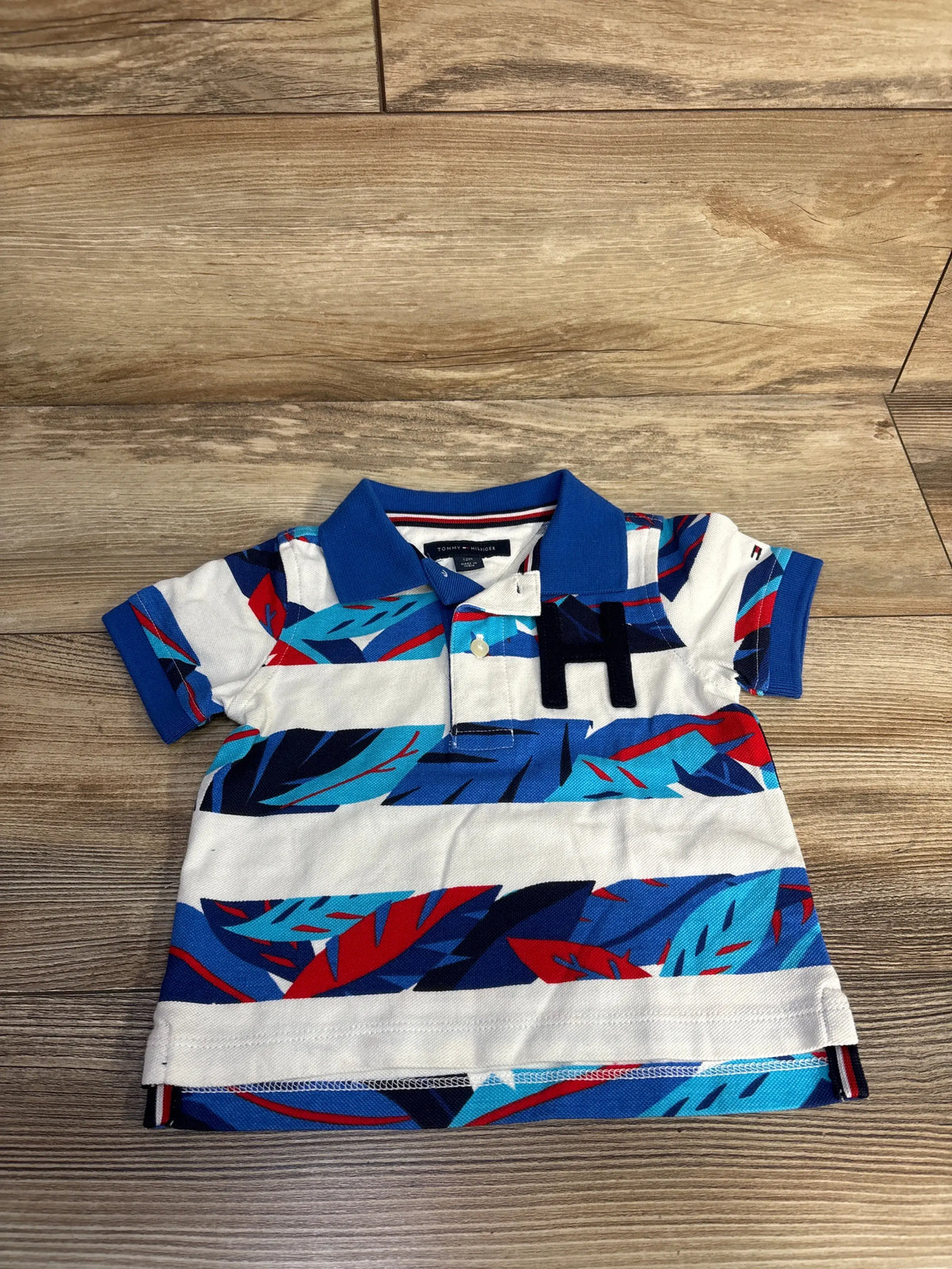 Front view of Tommy Hilfiger polo shirt in white, size 12 months, showcasing a blue and red pattern with button front on a wood floor
