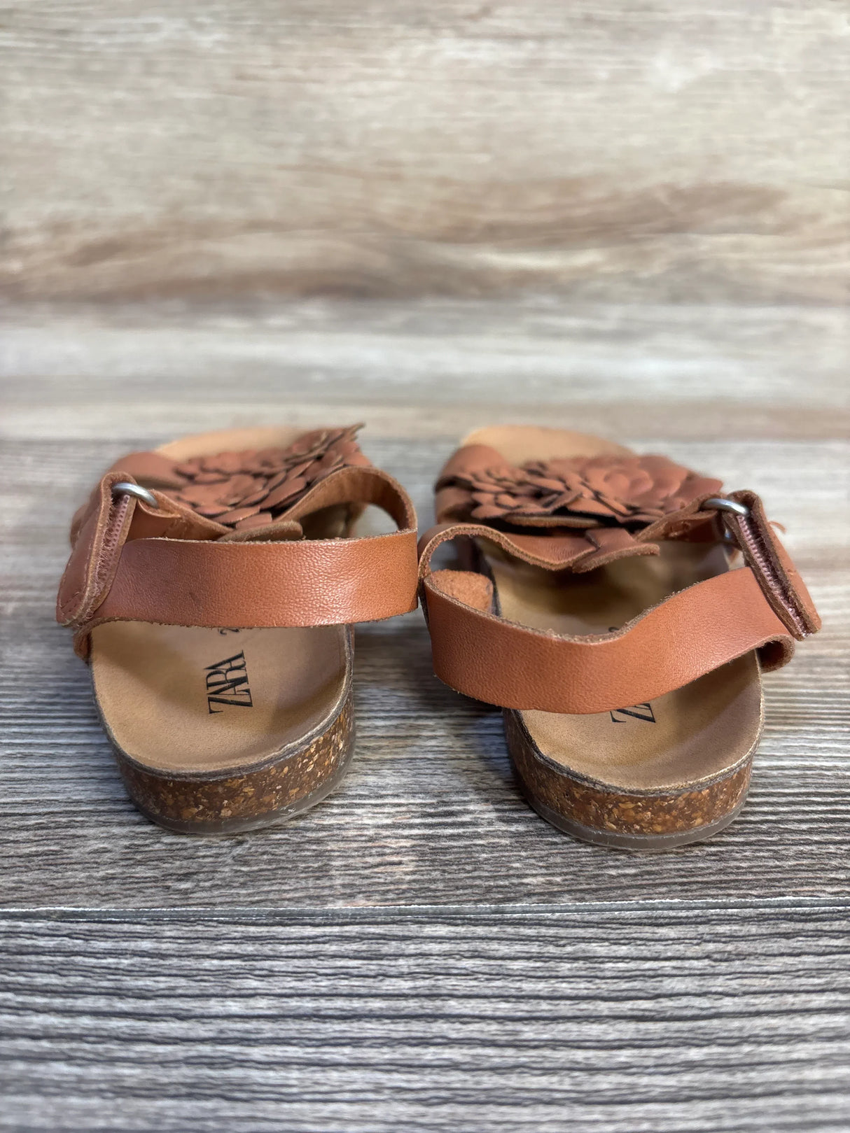 Brown toddler sandals with floral detail, cork soles, and ankle straps on wood surface
