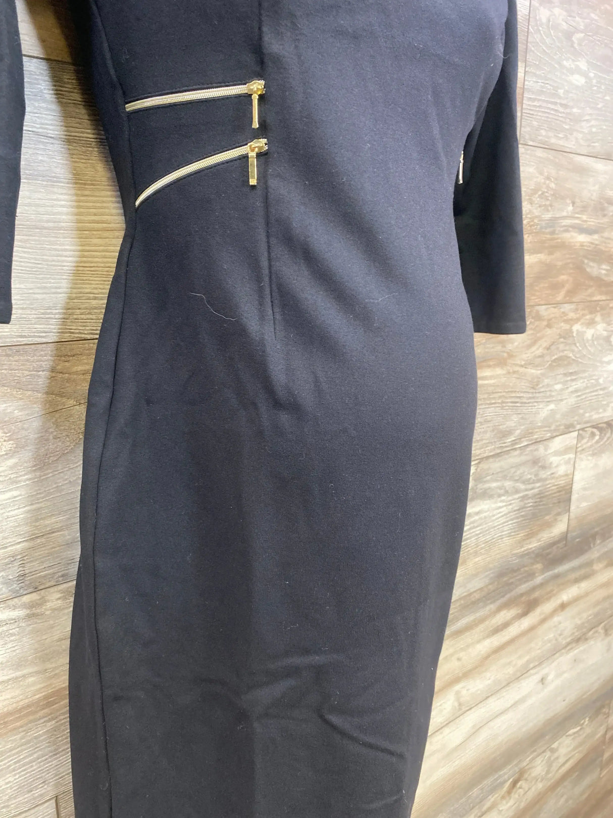 Front view of black Seraphine 3/4 sleeve maternity dress showcasing angled gold zipper accents with wood-paneled background