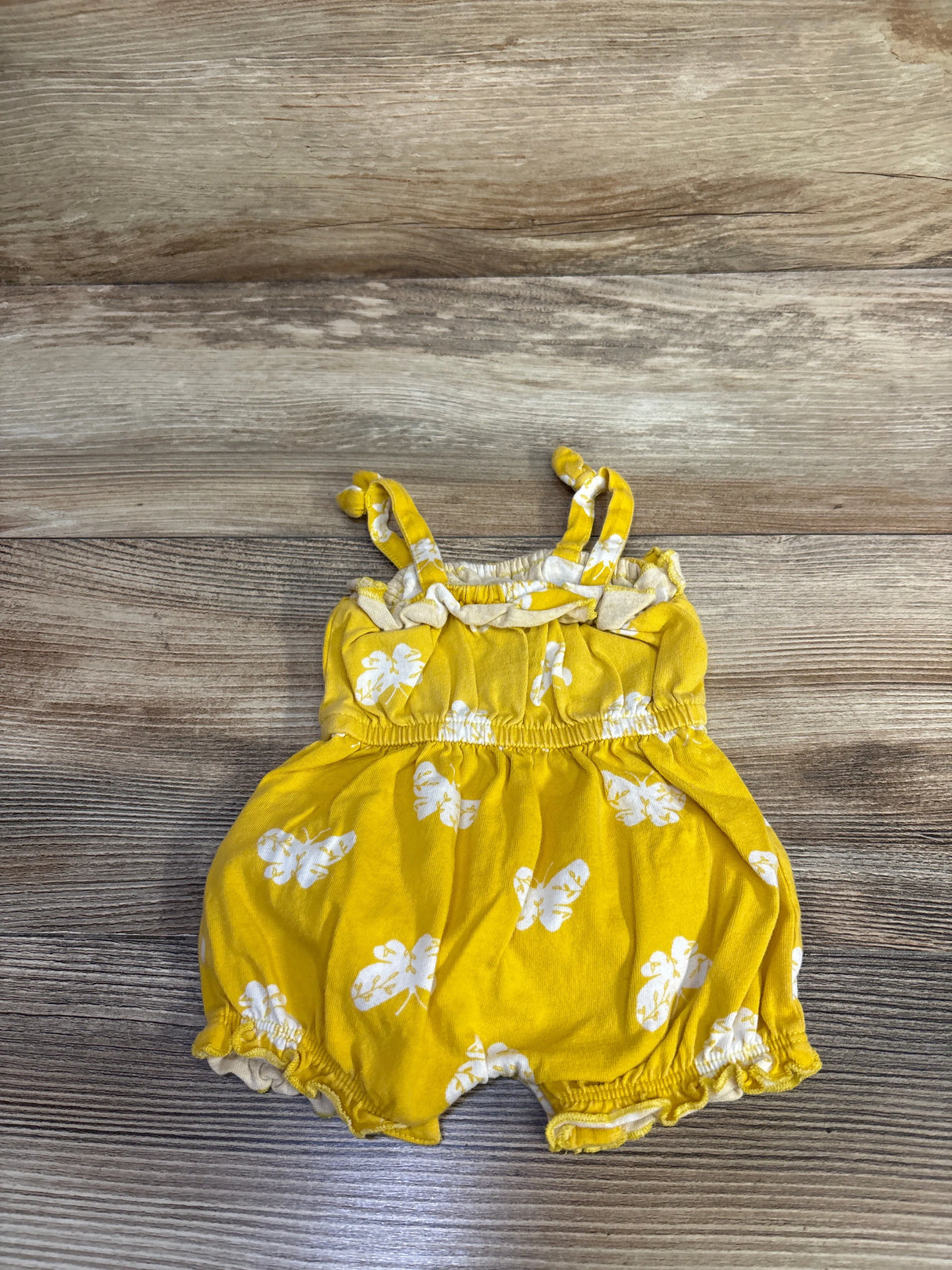 Flat lay newborn yellow shortie romper with white floral print, knotted straps, elastic waist, ruffled legs on wood floor