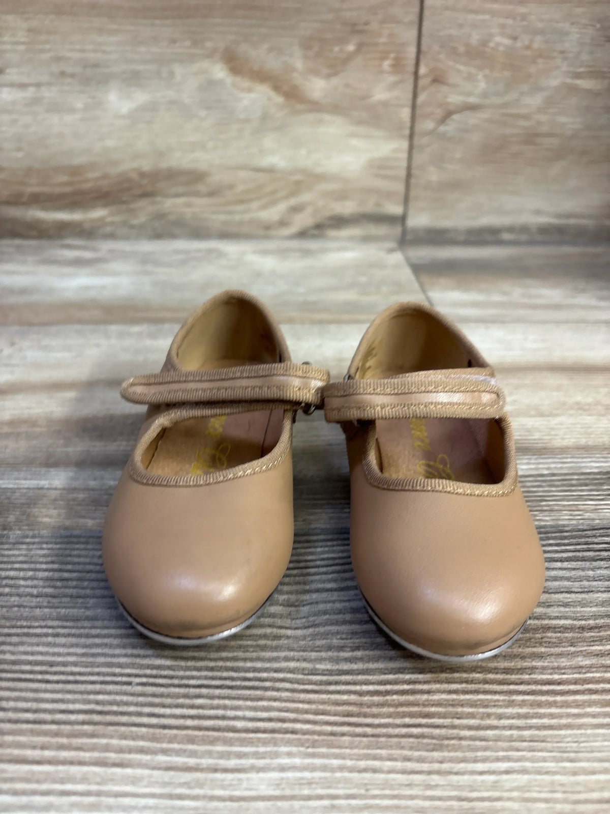Front view of tan tap shoes in size 7C showcasing wide straps and rounded toes on a wood-grain floor