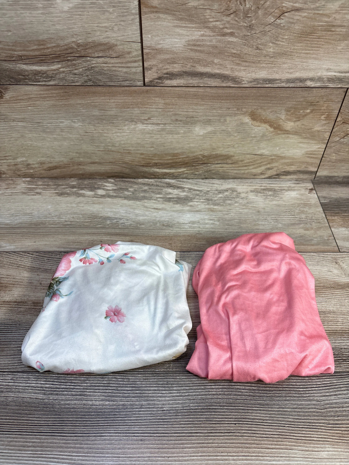 Folded view of two fitted bassinet sheets showcasing white floral print and solid pink design on a wood background