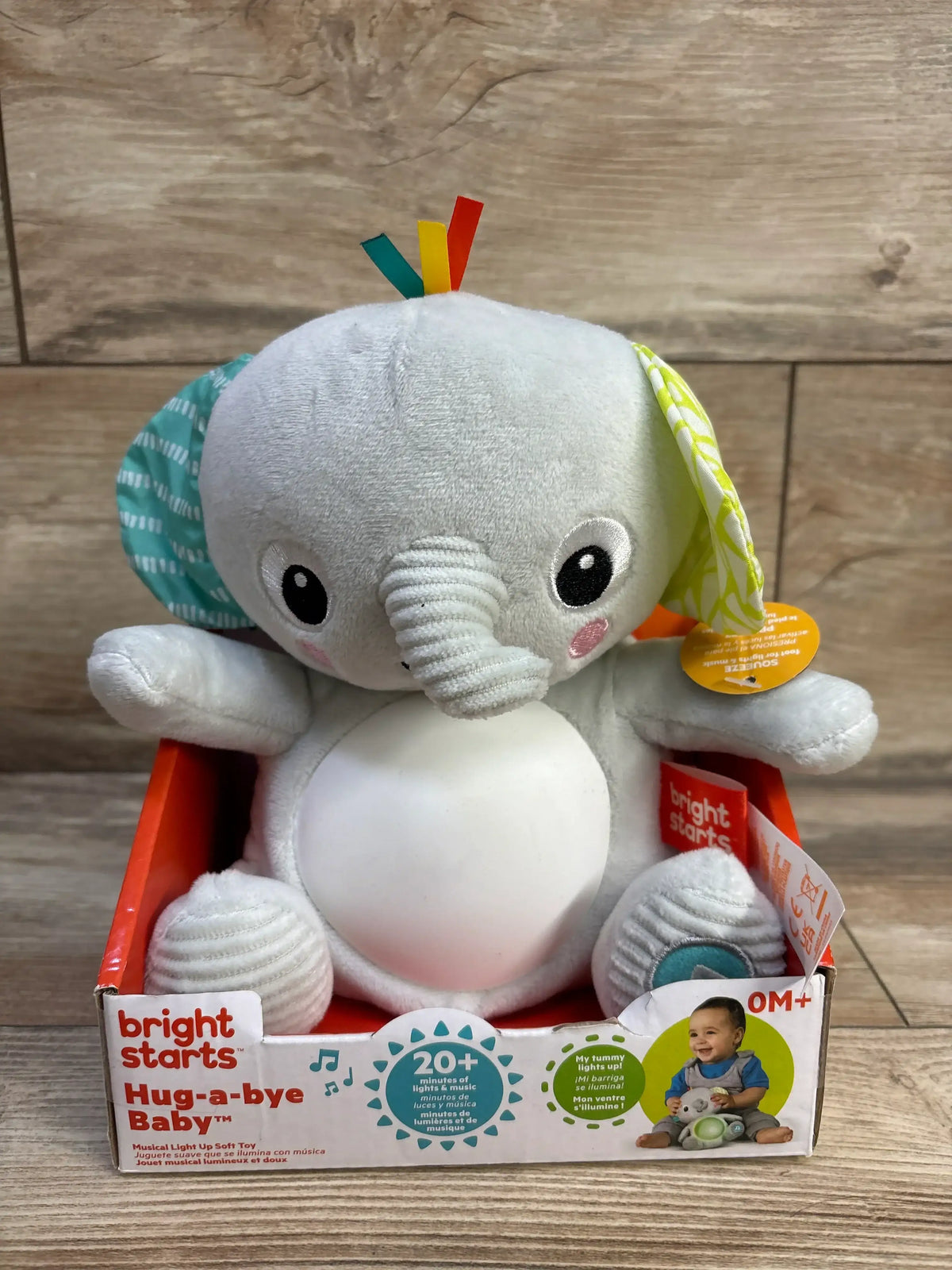 Front view of Hug-a-bye elephant plush with light-up tummy, patterned ears and ribbon tags in box on wood floor