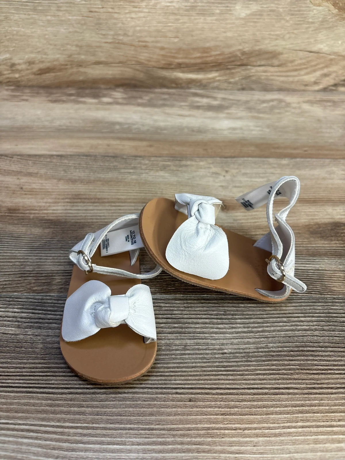 Top down view of white leather baby sandals showing knotted bows and ankle straps with gold buckles on a wood-grain background