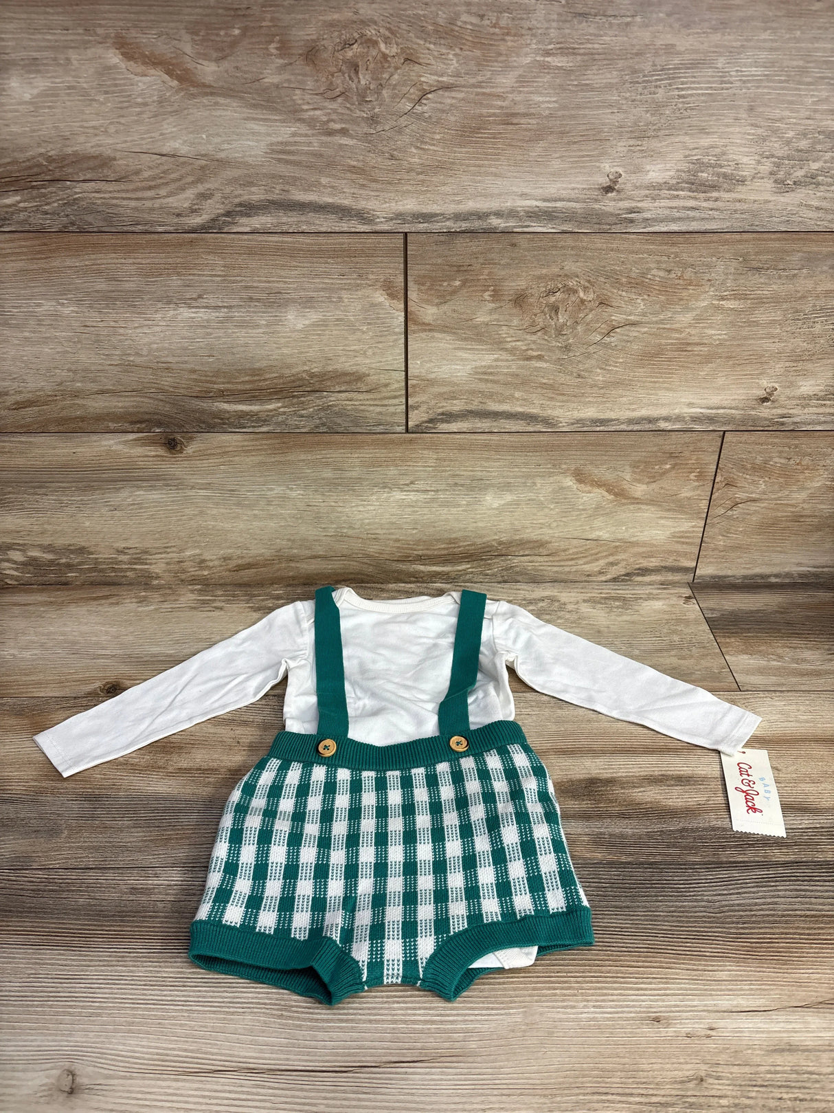White long-sleeve baby bodysuit with green and white checkered knit suspender shorts on wooden floor