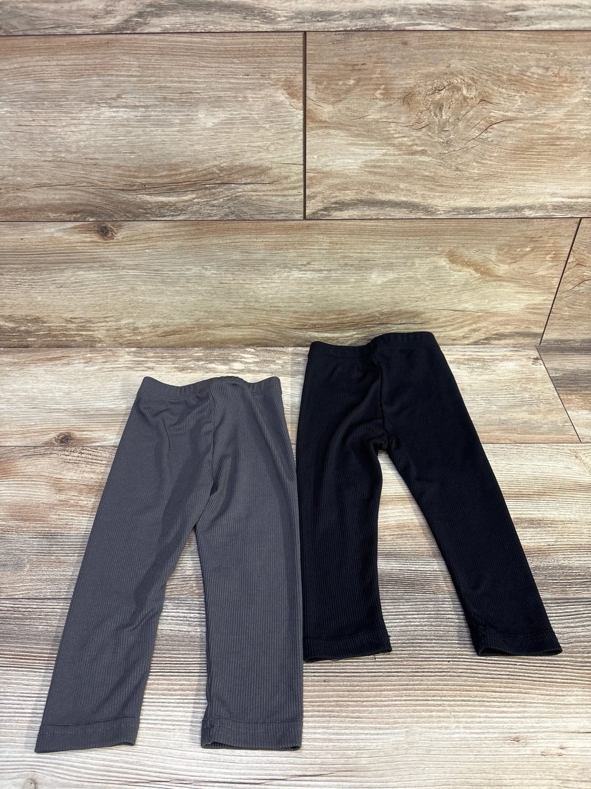 Flat lay of two ribbed baby leggings showing elastic waist and ribbed texture, black and charcoal on wood background