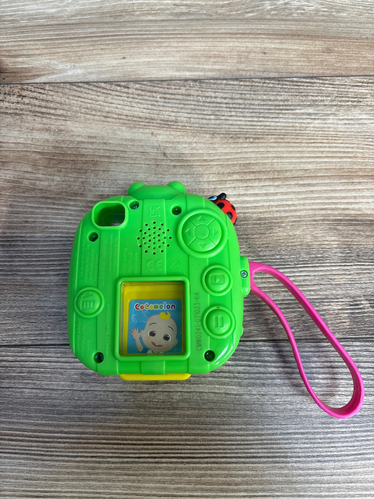 Green CoComelon kids toy camera with control buttons and pink wrist strap on wood surface
