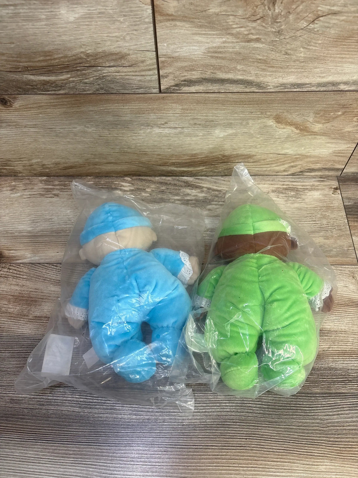 Top-down view of two Lakeshore washable cuddly baby dolls, one blue and one green, in plastic bags on wood floor