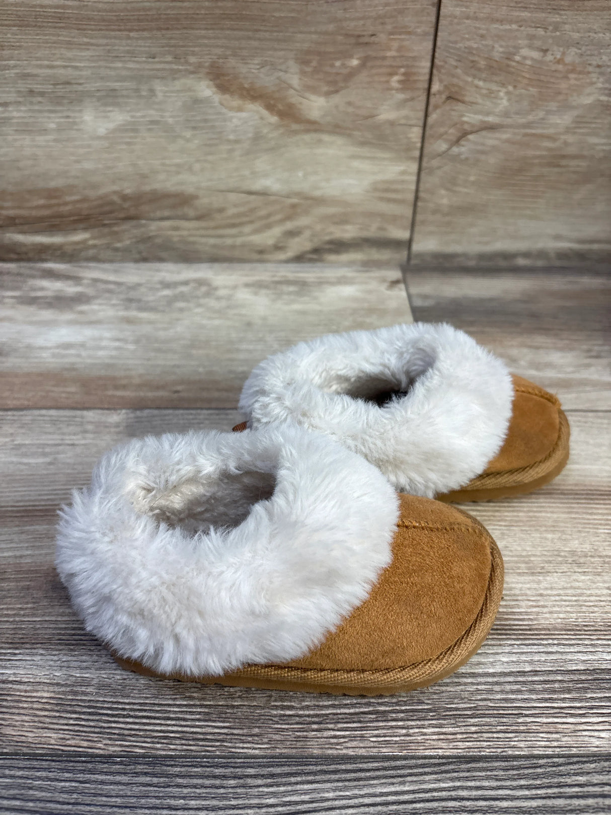 Angled top view of brown baby slippers with white faux-fur cuffs showcasing plush lining and suede upper on wood floor