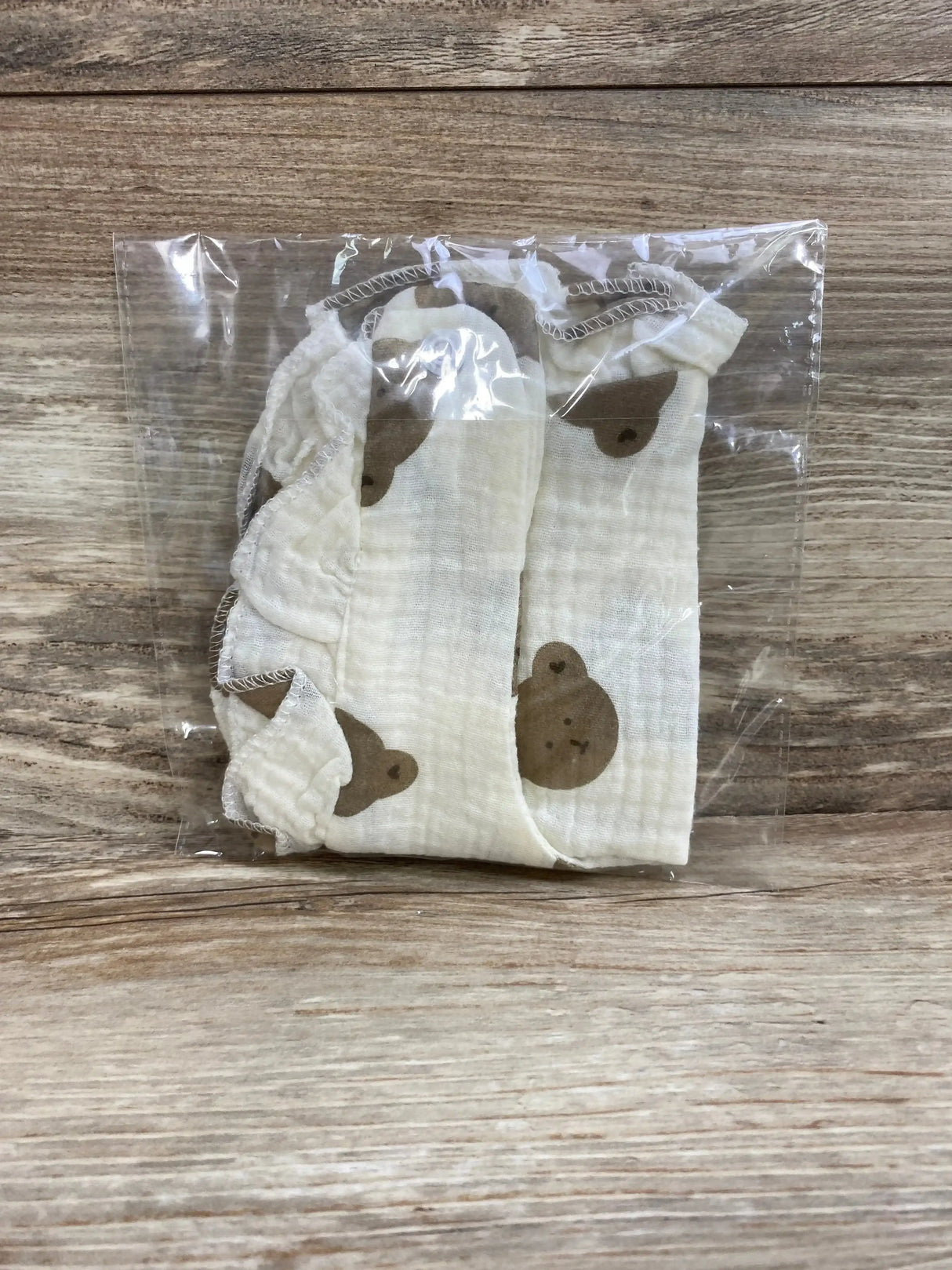 Top-down view of folded ruffle cotton baby bib showcasing cream color and brown bear print with ruffled edge in clear plastic packaging on wood background