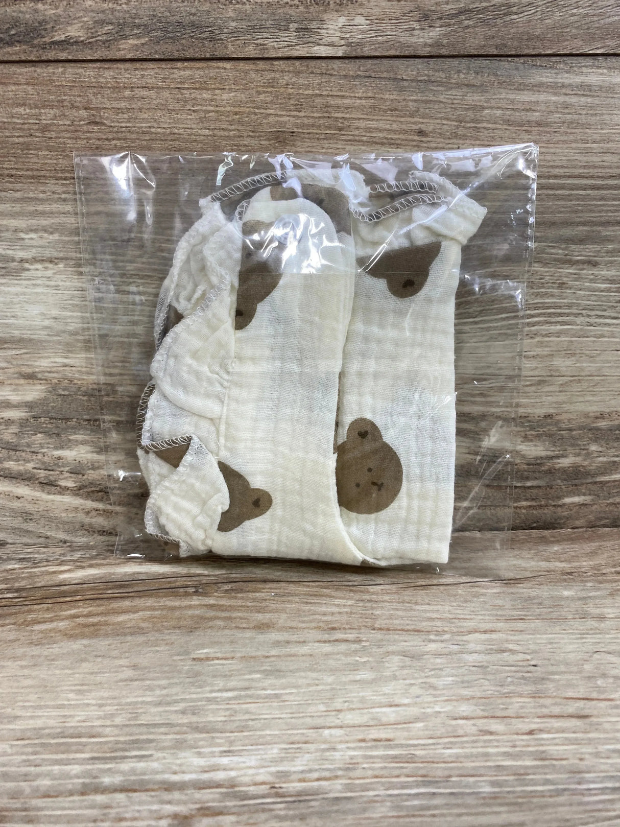 Top-down view of folded ruffle cotton baby bib showcasing cream color and brown bear print with ruffled edge in clear plastic packaging on wood background