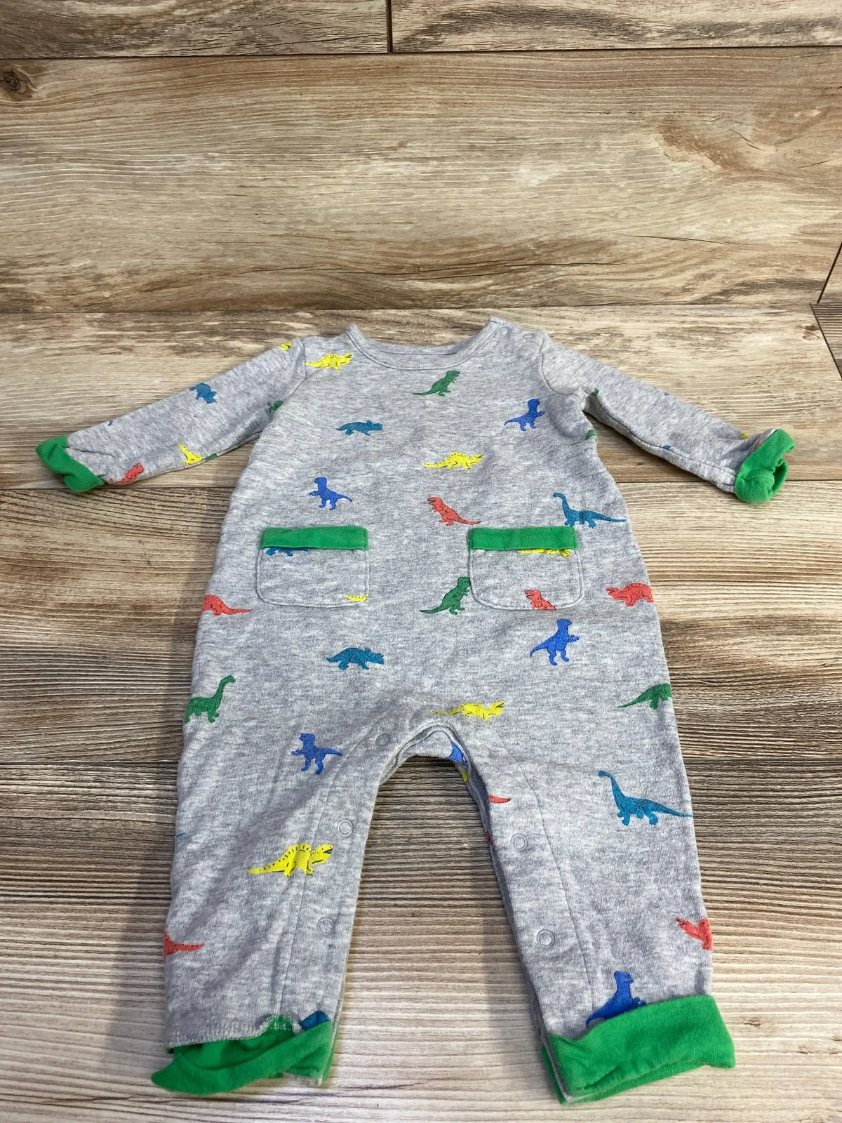 Grey baby onesie with colorful dinosaur prints and green cuffs and pocket trim on wood floor