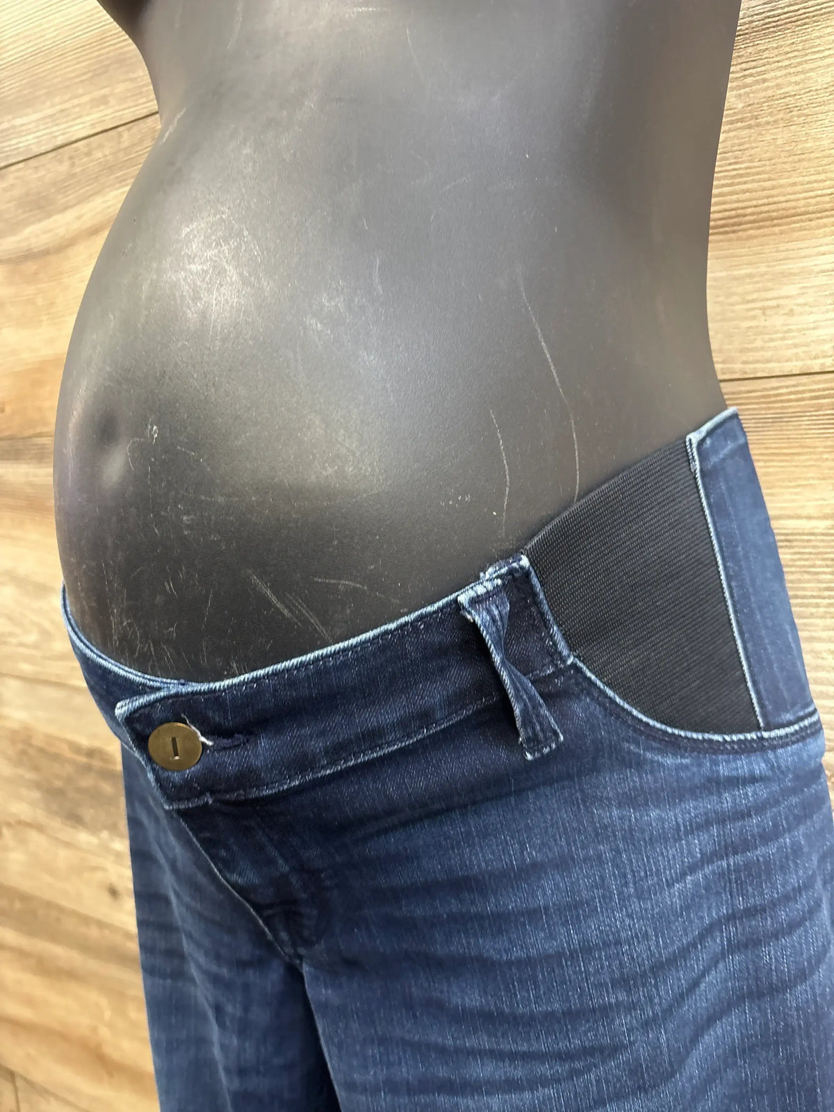 Side view of navy Isabel maternity skinny jeans showing black elastic side panel and low-rise button waist on mannequin against wood backdrop