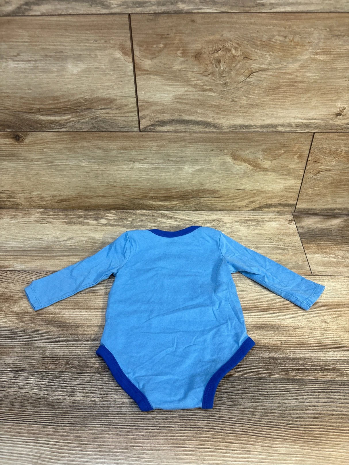 Back view flat lay of blue Nike long-sleeve baby bodysuit (6 months) showing contrast dark-blue trim at neckline and leg openings on wood floor background