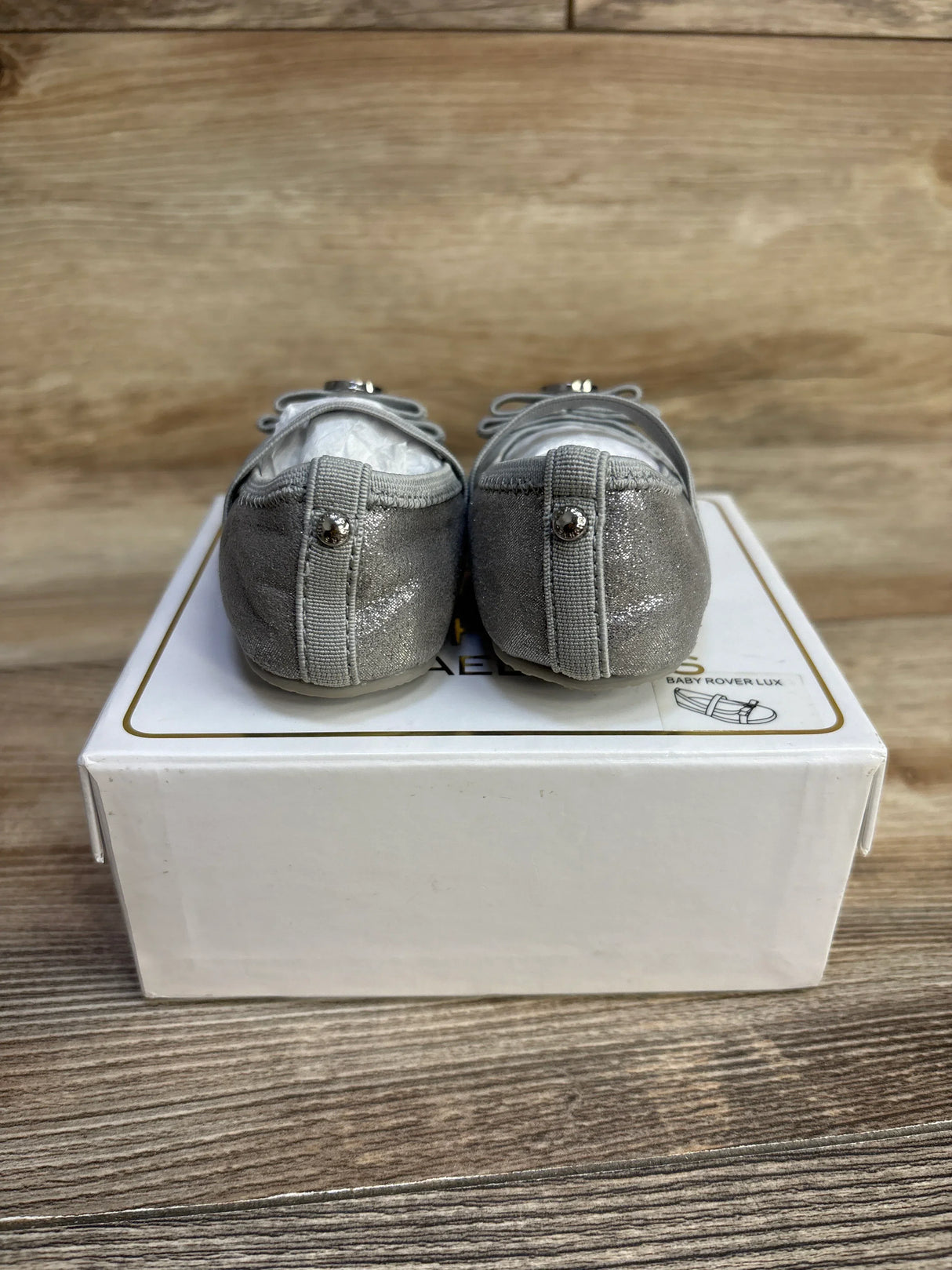 Rear view of Michael Kors Baby Rover Lux baby shoes showcasing glittery silver fabric and heel stud detail with white box on wood background