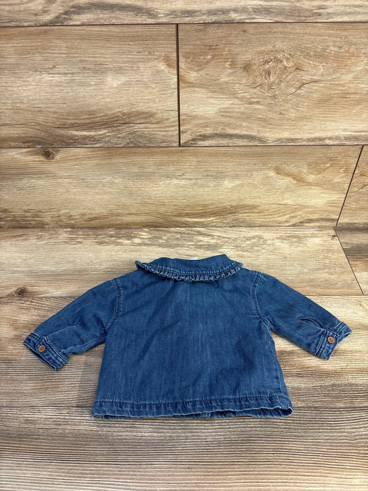 Back view of Next blue denim jacket, size 0-3 months, showcasing ruffled collar and buttoned cuffs on woodgrain surface