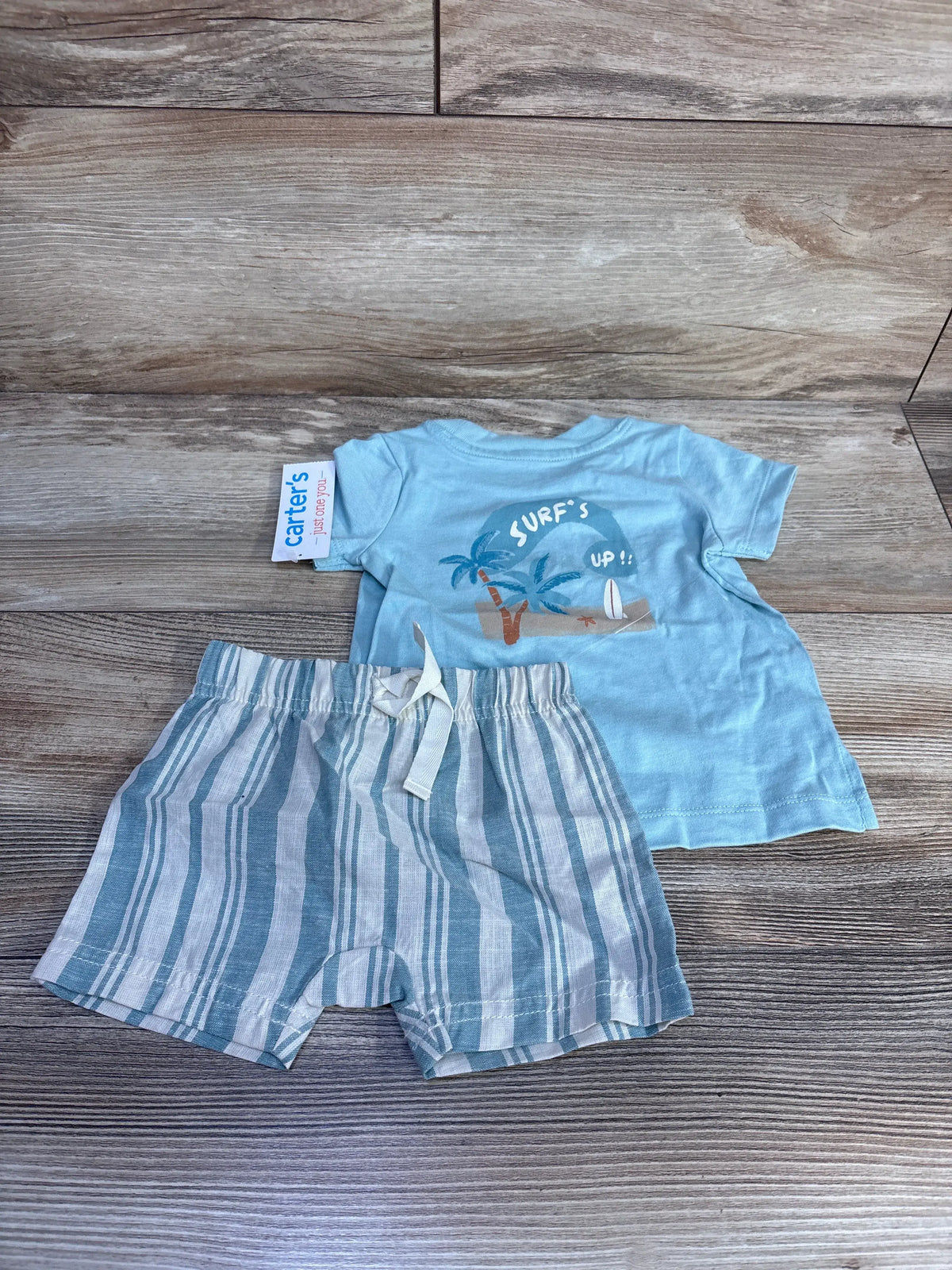 Flat lay of blue Just One You two-piece baby outfit set with surf-themed shirt and striped shorts on wood grain