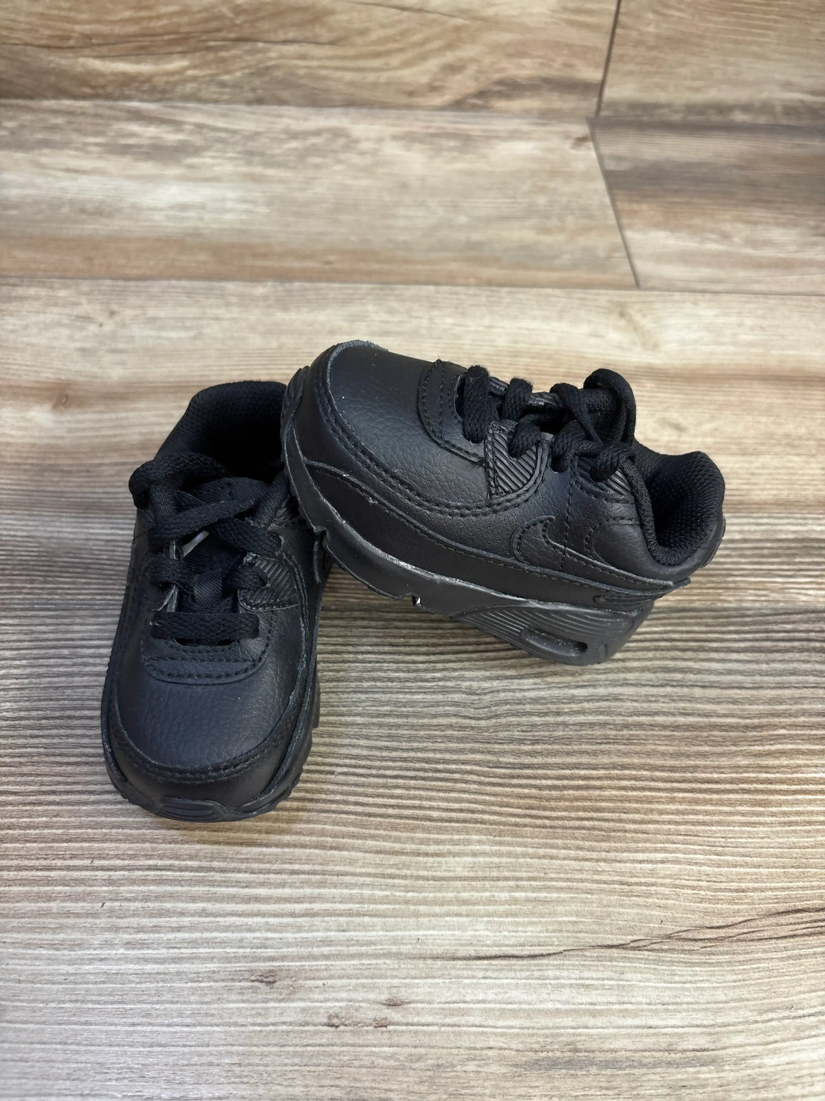 Angled view of Nike Air Max 90 TD toddler sneakers showcasing all-black design on a wood-look floor