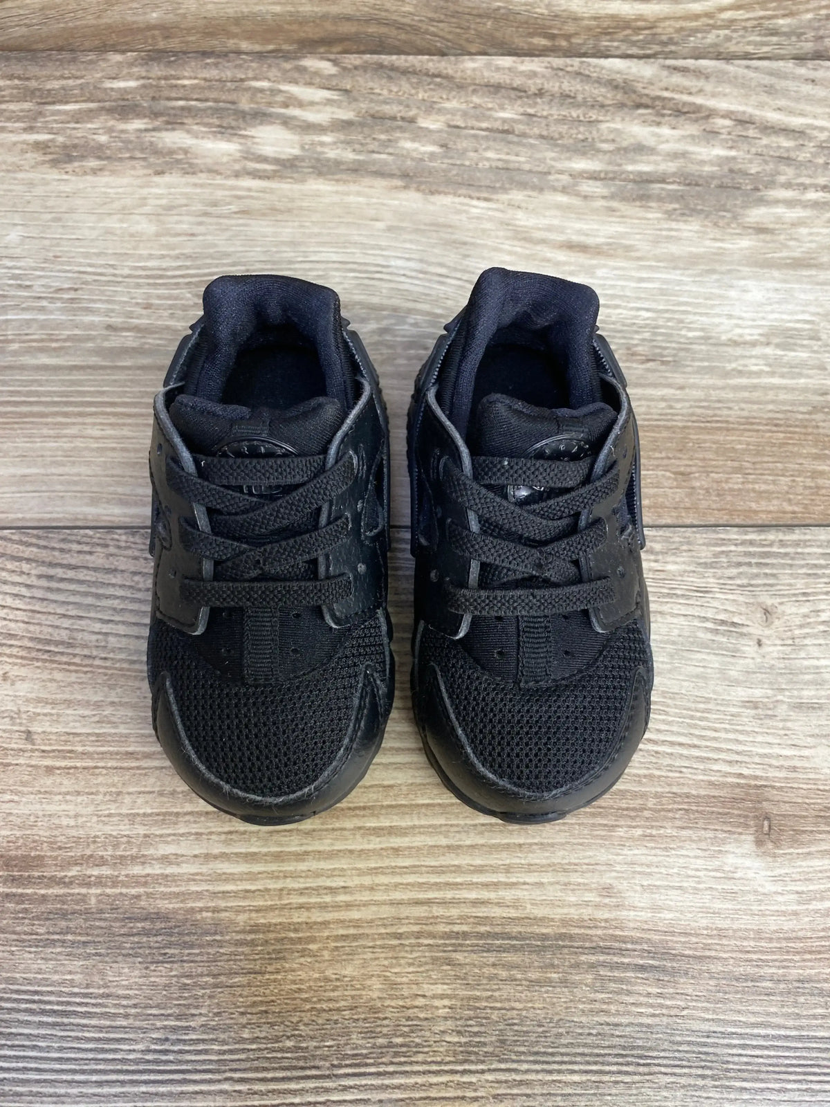 Top view of Nike Air Huarache Run toddler sneakers, triple black, showing mesh toe, laces and Velcro strap on wood floor