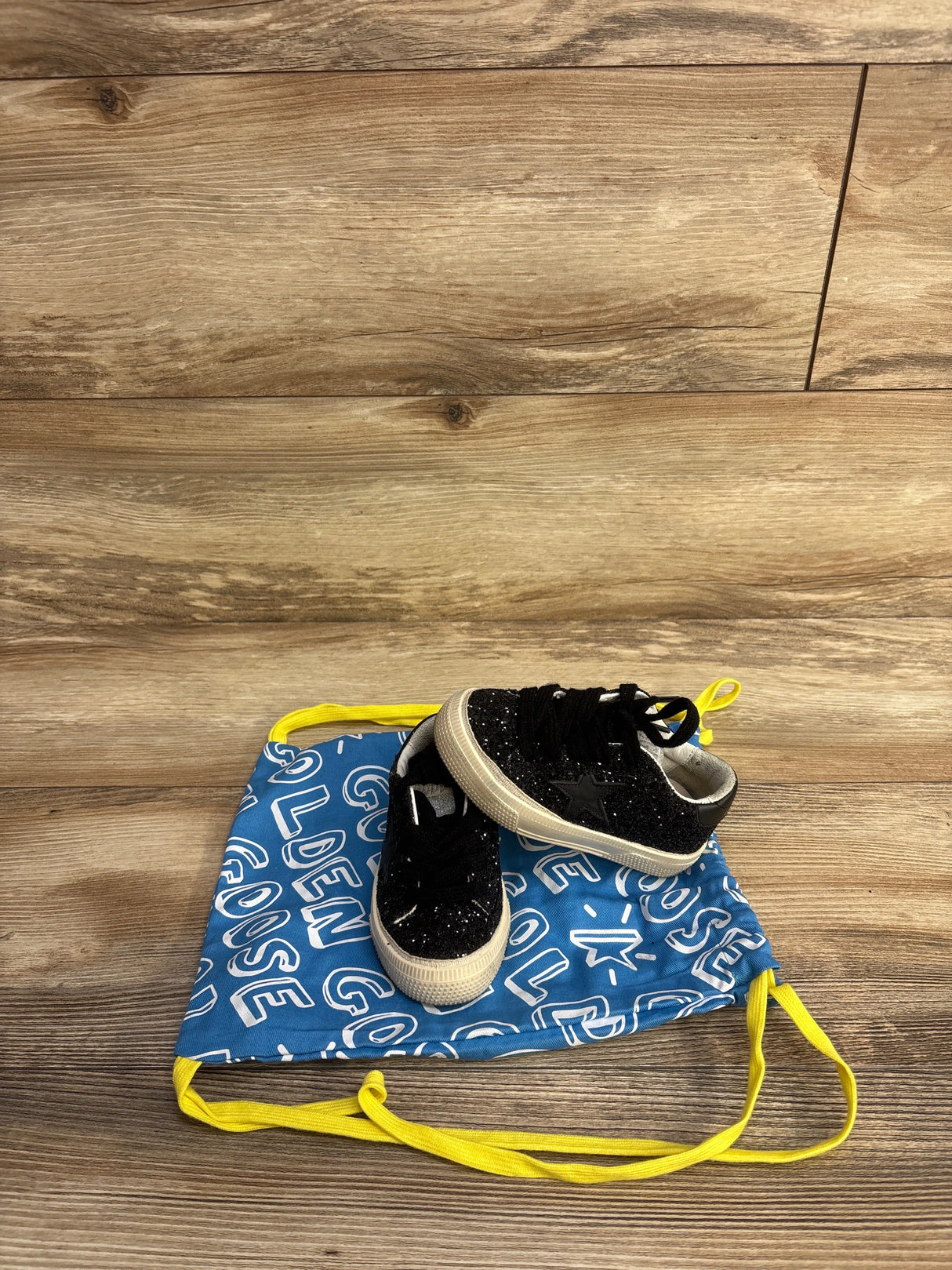 Black glitter toddler sneakers on a blue Golden Goose dust bag with yellow drawstrings on wood floor