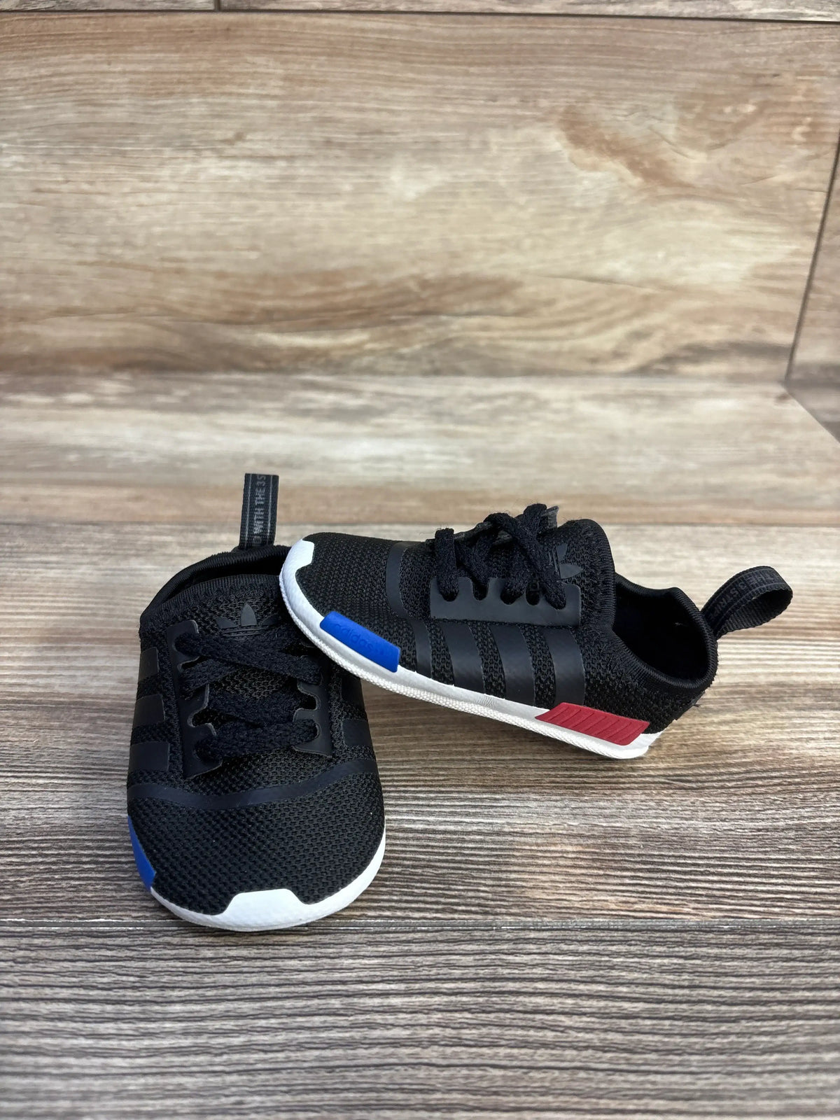 Angled view of black Adidas NMD Soft Sole Sneakers showcasing elastic lace closures and white soles on a wood-look floor