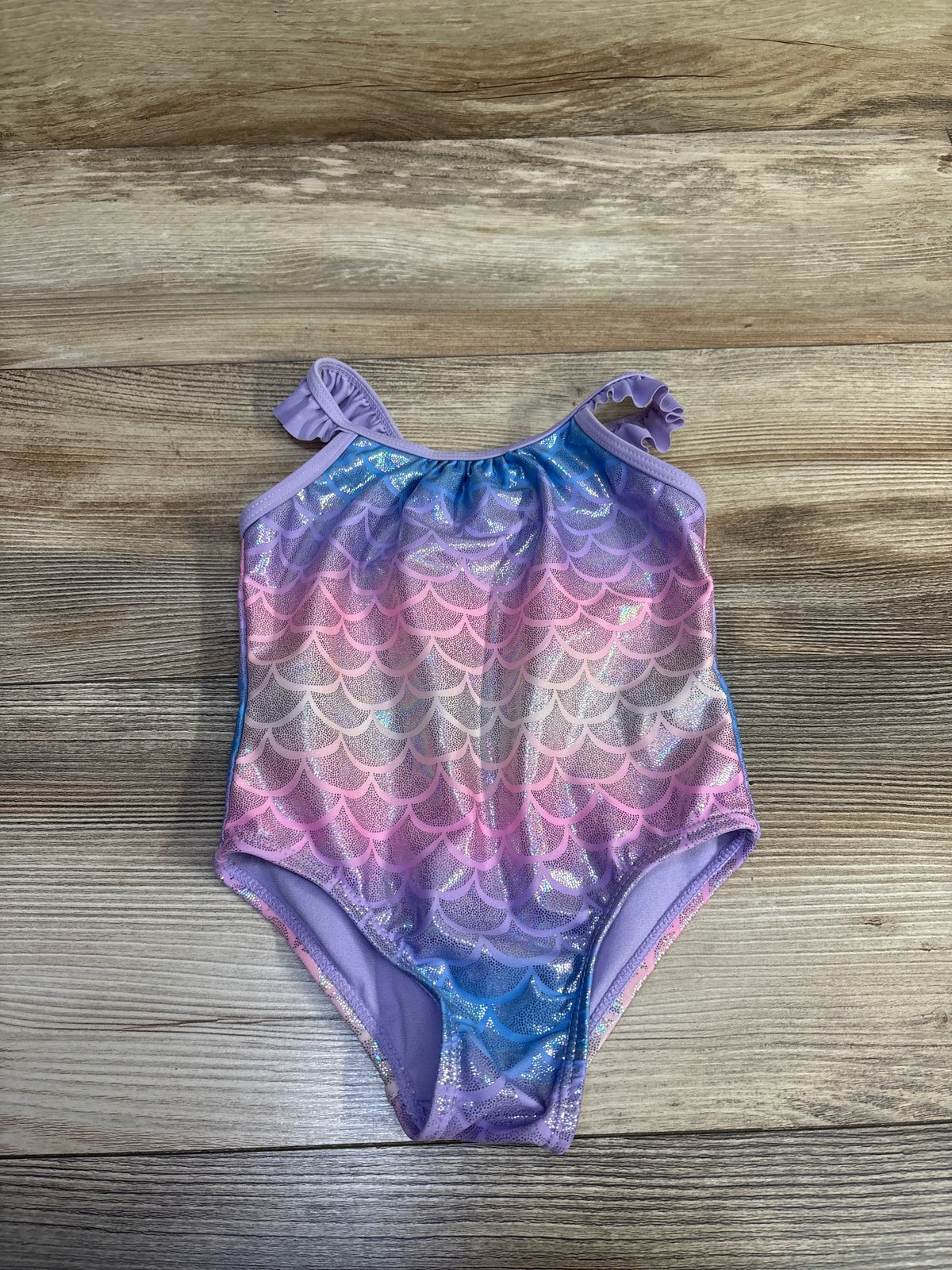 Front view of a purple Sol Borne one-piece mermaid swimsuit with shimmering scallop pattern and tie shoulder straps on a wood-grain surface