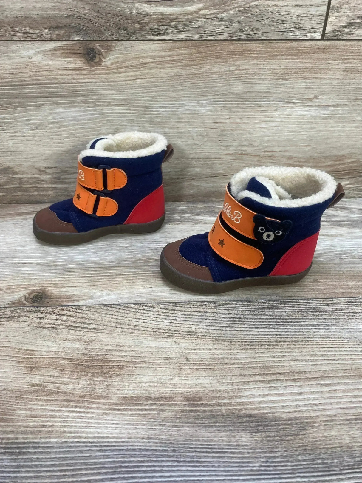 Three-quarter view of toddler winter boots in navy, orange, and red with faux-fur lining, velcro straps, bear patch on wood background