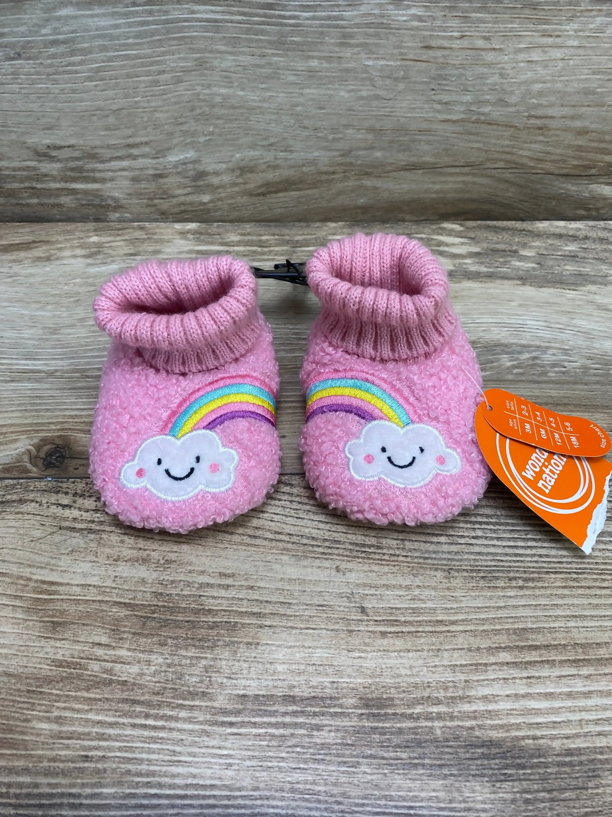 Front view of pink baby bootie slippers showing embroidered smiling cloud and rainbow patch with ribbed cuff on wood background