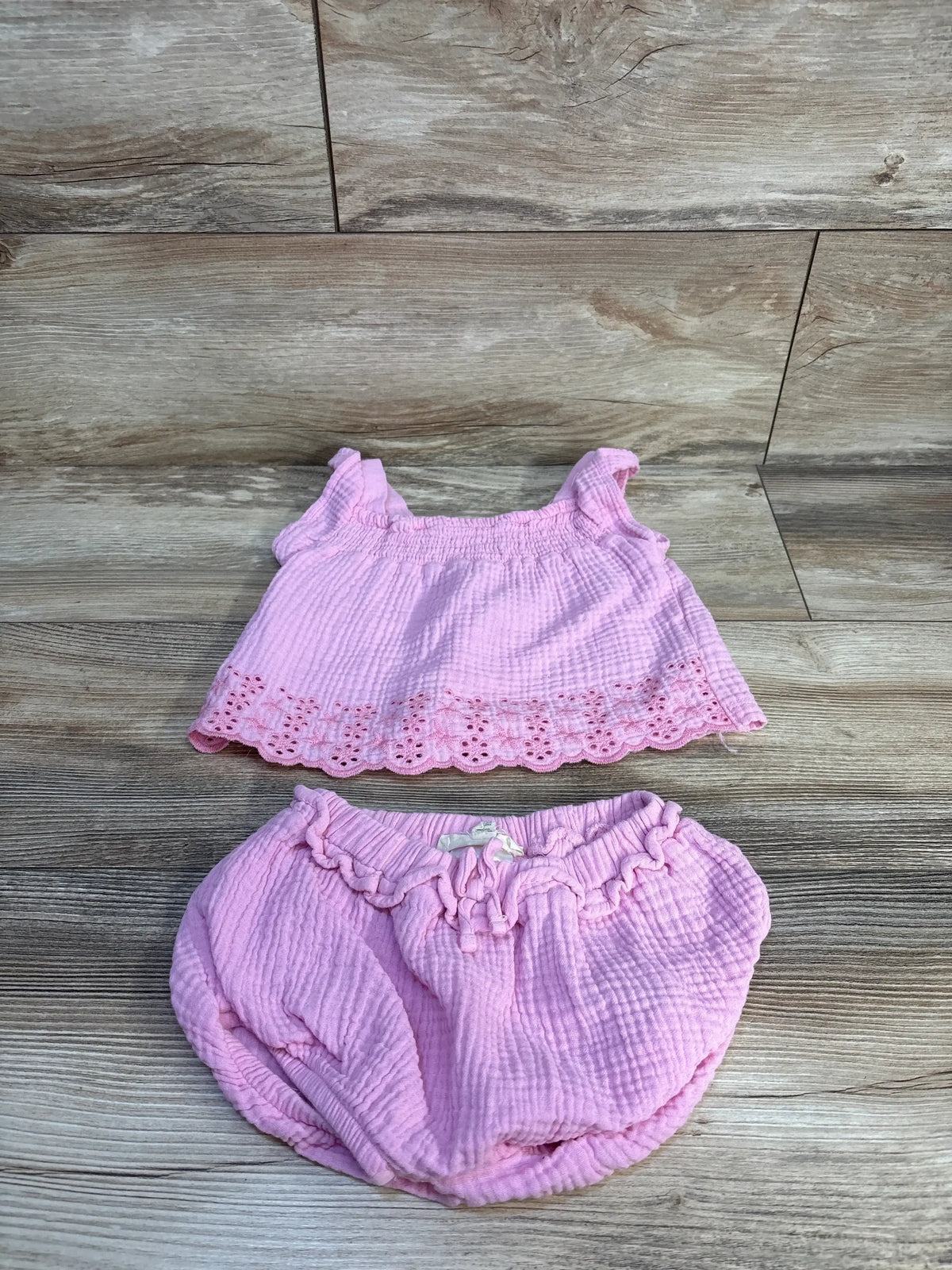 Pink baby girl two-piece outfit with eyelet trim top and ruffled shorts, laid on wood floor