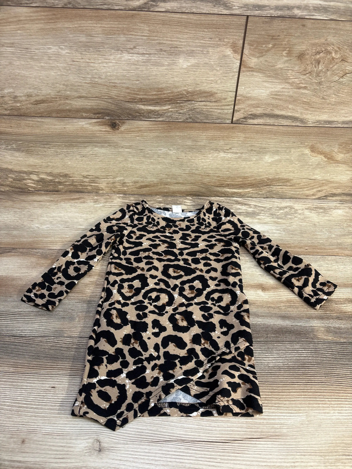 Flat lay of beige leopard-print long-sleeve toddler dress showcasing tulip wrap hem and crew neckline with wood floor background