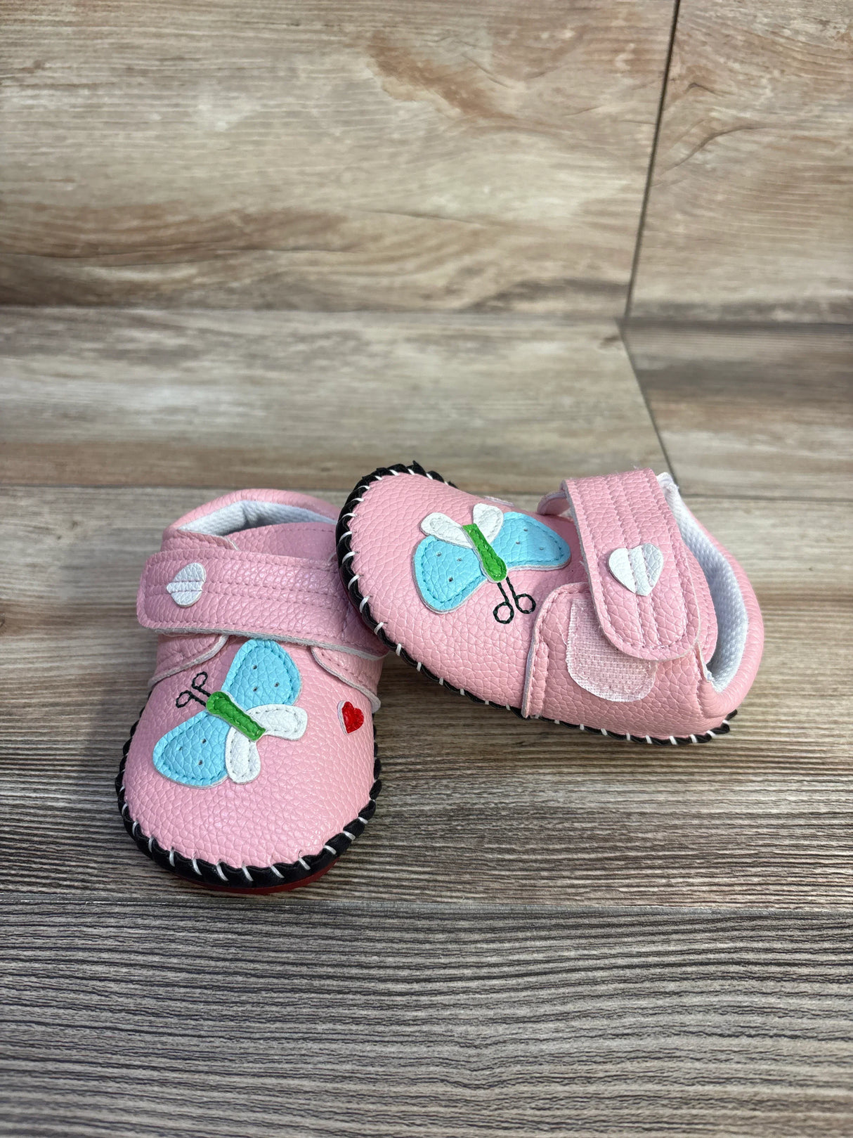 Angled front view of pink PU baby sneakers with blue butterfly and heart patches and black stitched sole on wood floor