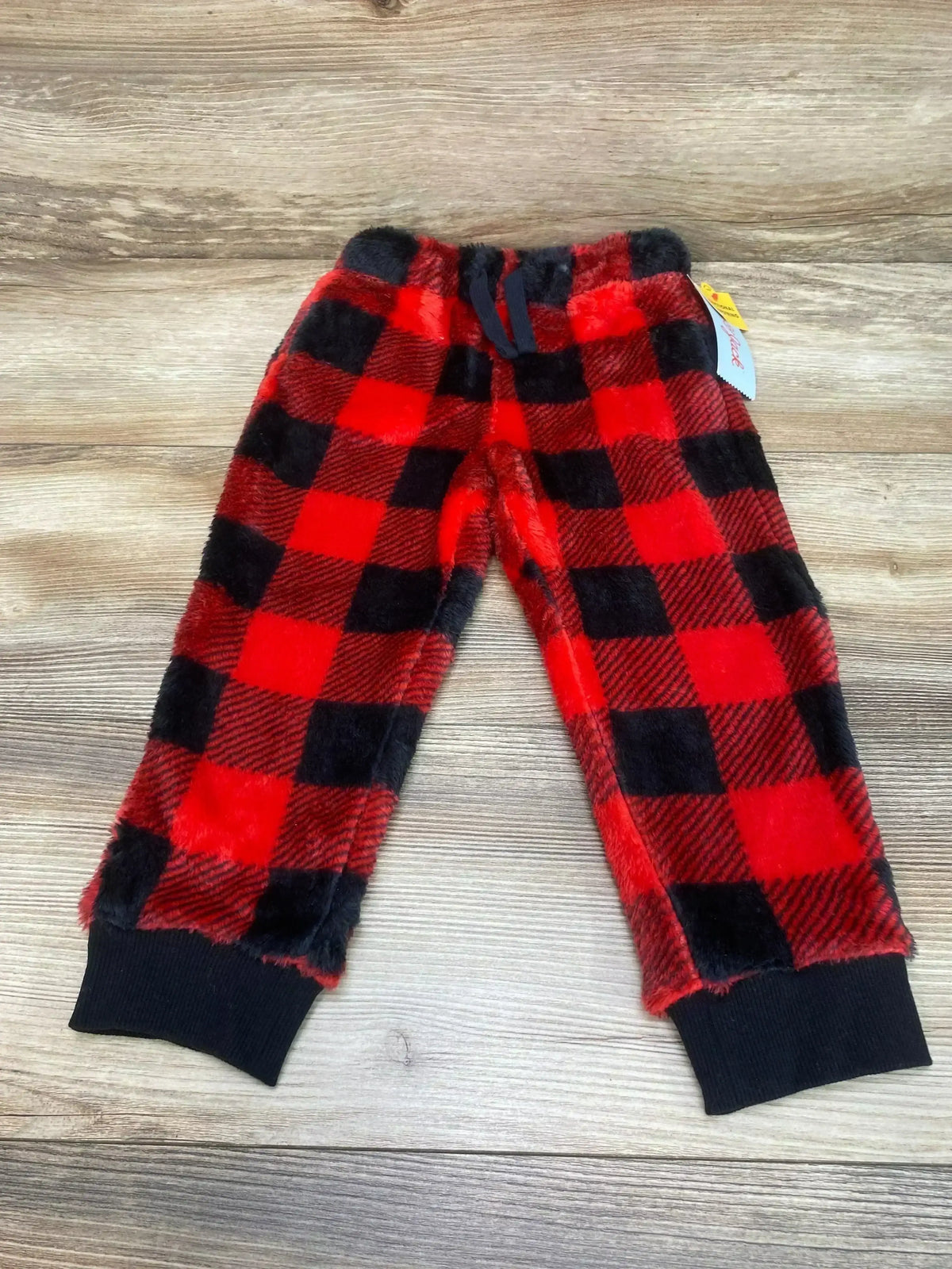 Flat-lay of toddler red and black buffalo-check cozy fleece pants with elastic waist, navy drawstring and black ribbed cuffs on wood background
