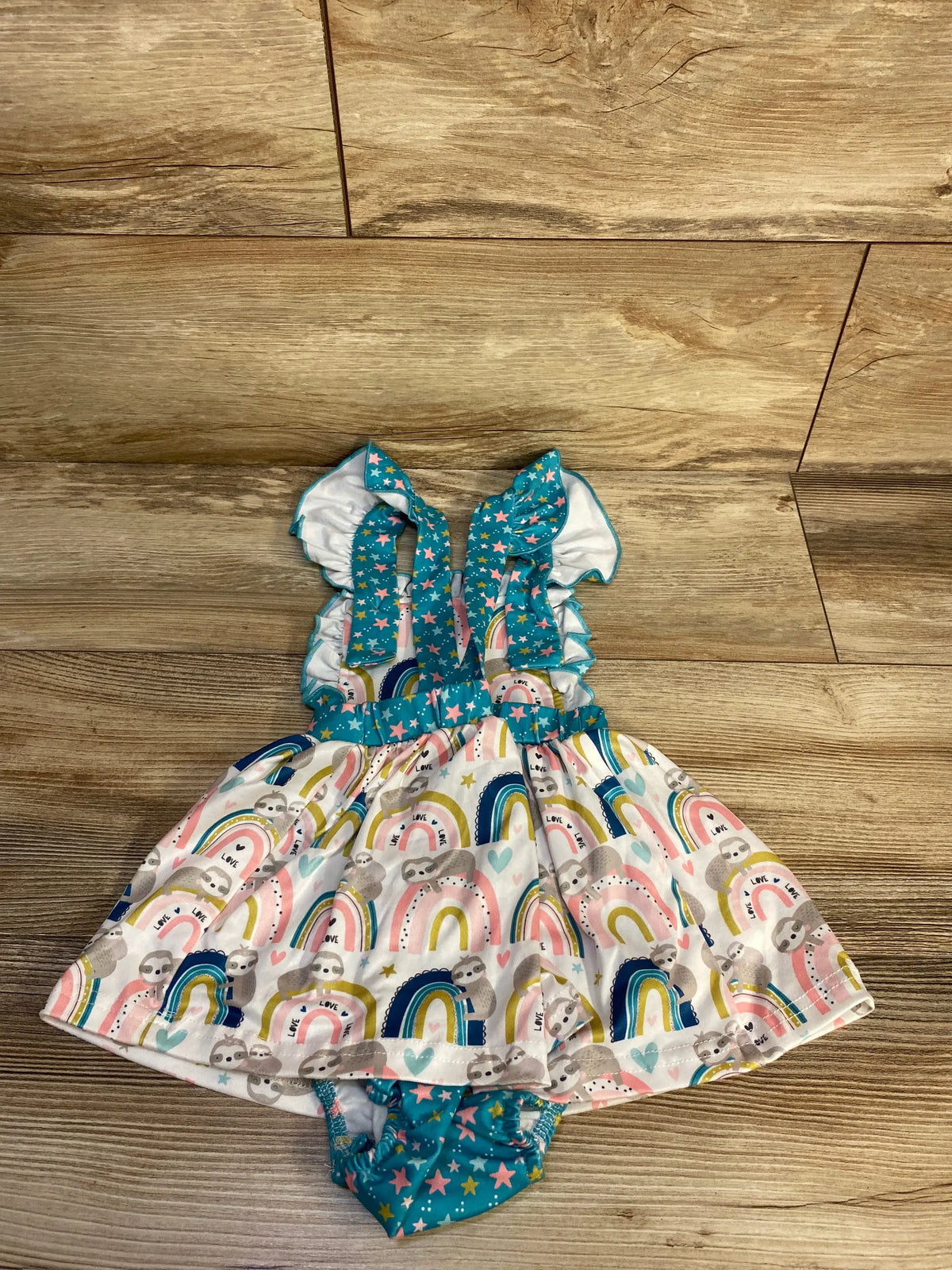 Flat lay of Pete + Lucy sloth bodysuit dress showcasing colorful sloth and rainbow prints on wood flooring