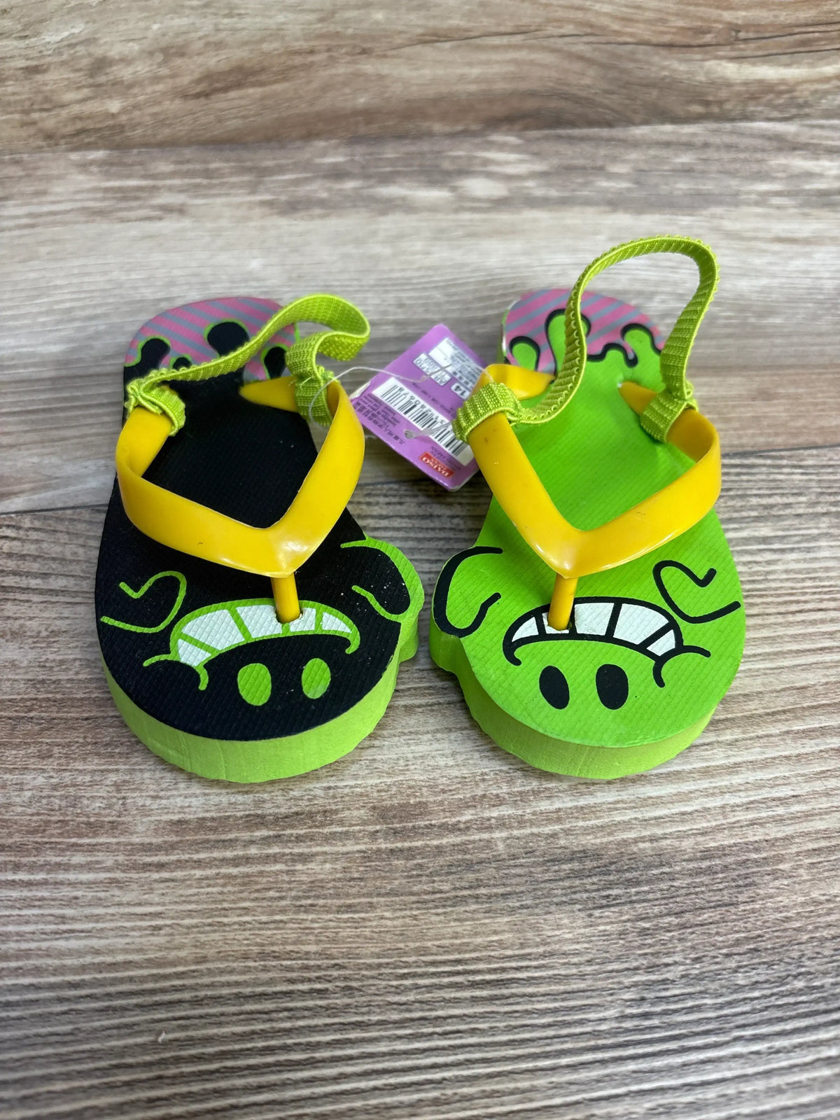 Top-down view of toddler flip-flops with lime green and black foam soles, yellow straps, and cartoon face prints