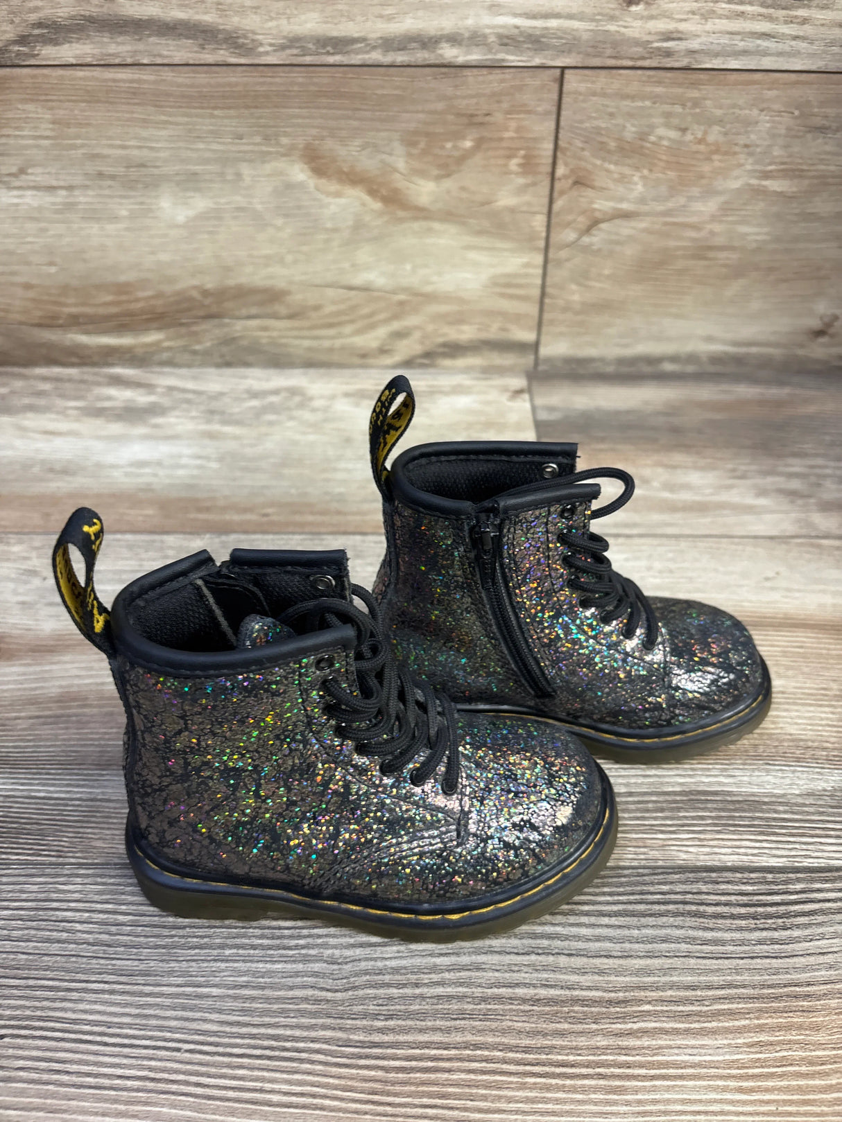 Three-quarter view of Dr. Martens 1460 Crinkle Metallic lace-up boots showcasing black crinkle metallic glitter finish, side zipper, yellow heel loop, and chunky sole on wood floor