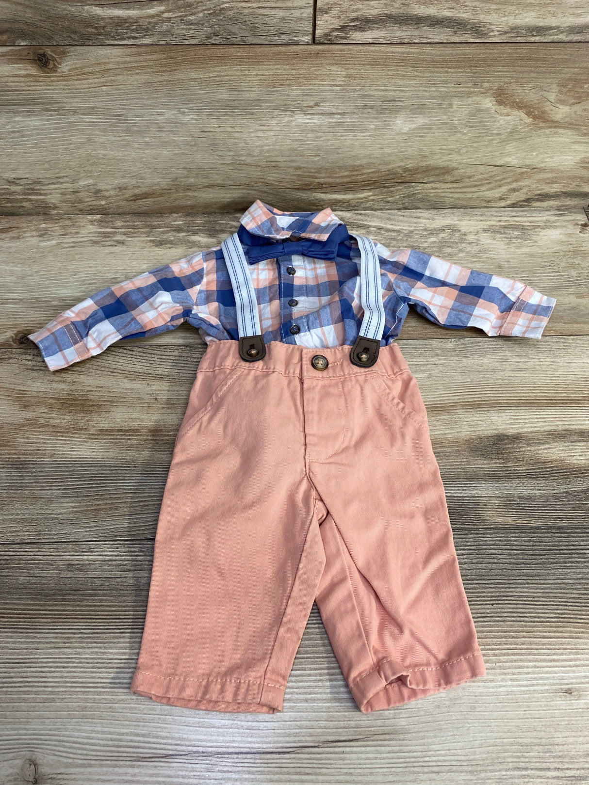 Baby boy outfit with pink pants, plaid shirt, suspenders, and blue bow tie on wood background