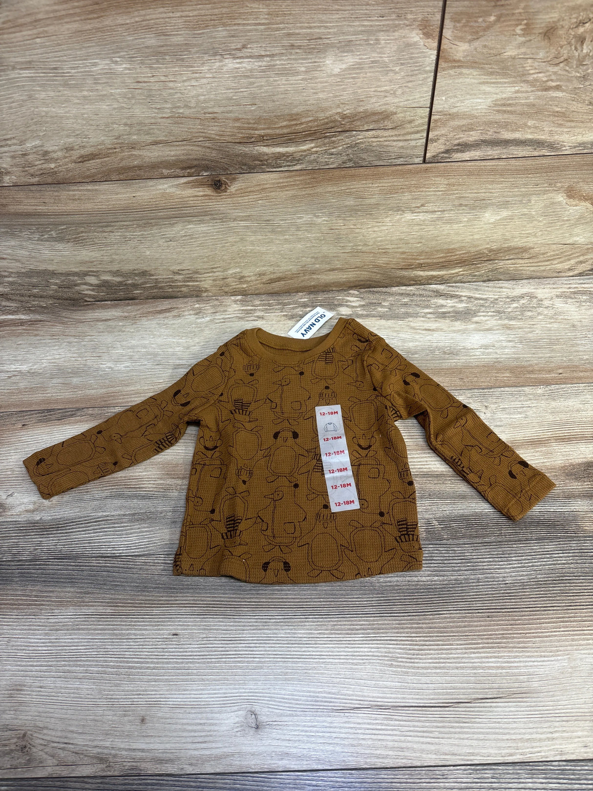 Brown long-sleeve toddler shirt with black animal illustrations and size tag 12-18M on wooden floor