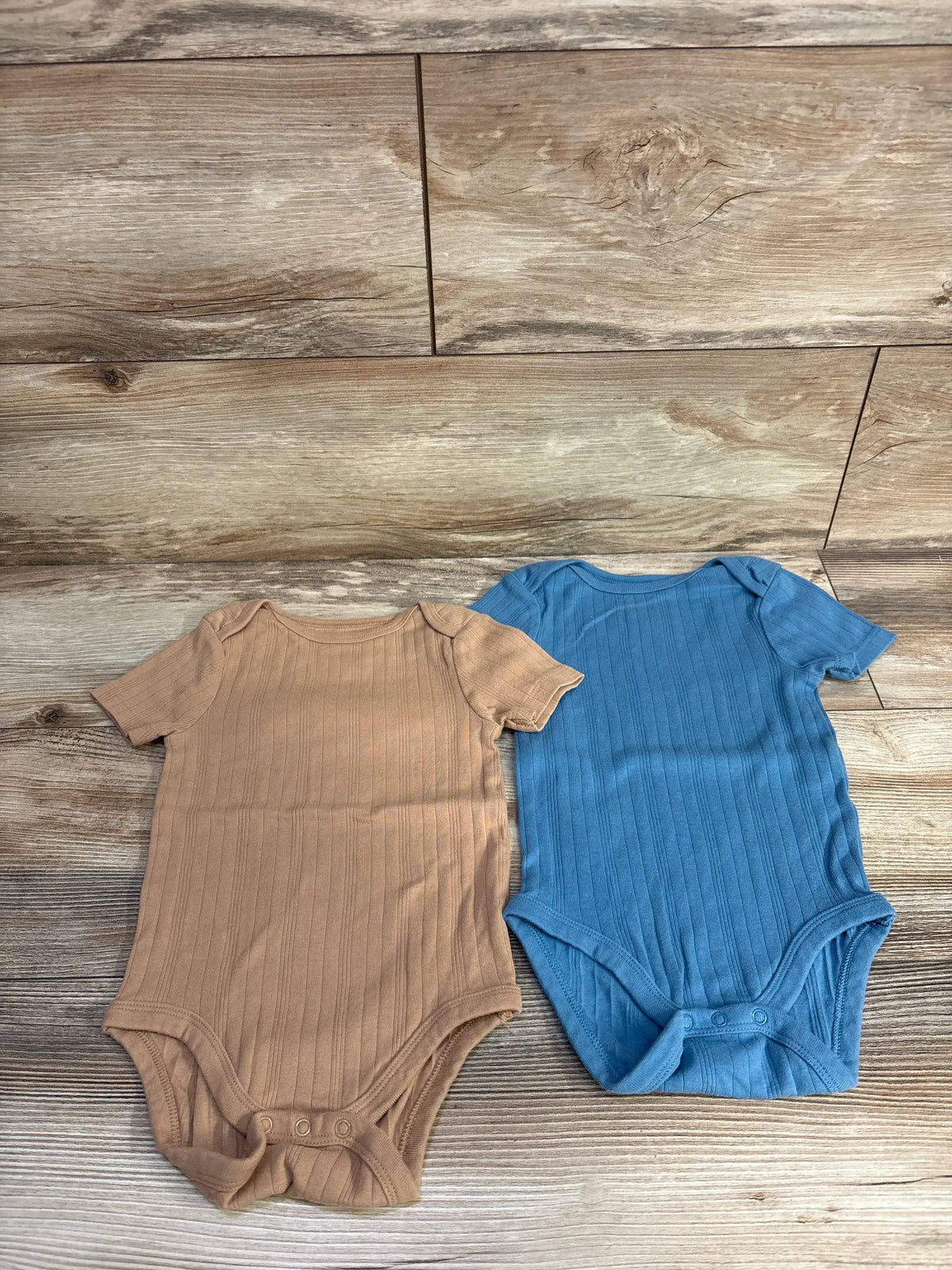Top view of Cloud Island two-pack ribbed bodysuits in blue with short sleeves and snap closures on a wood-grain background