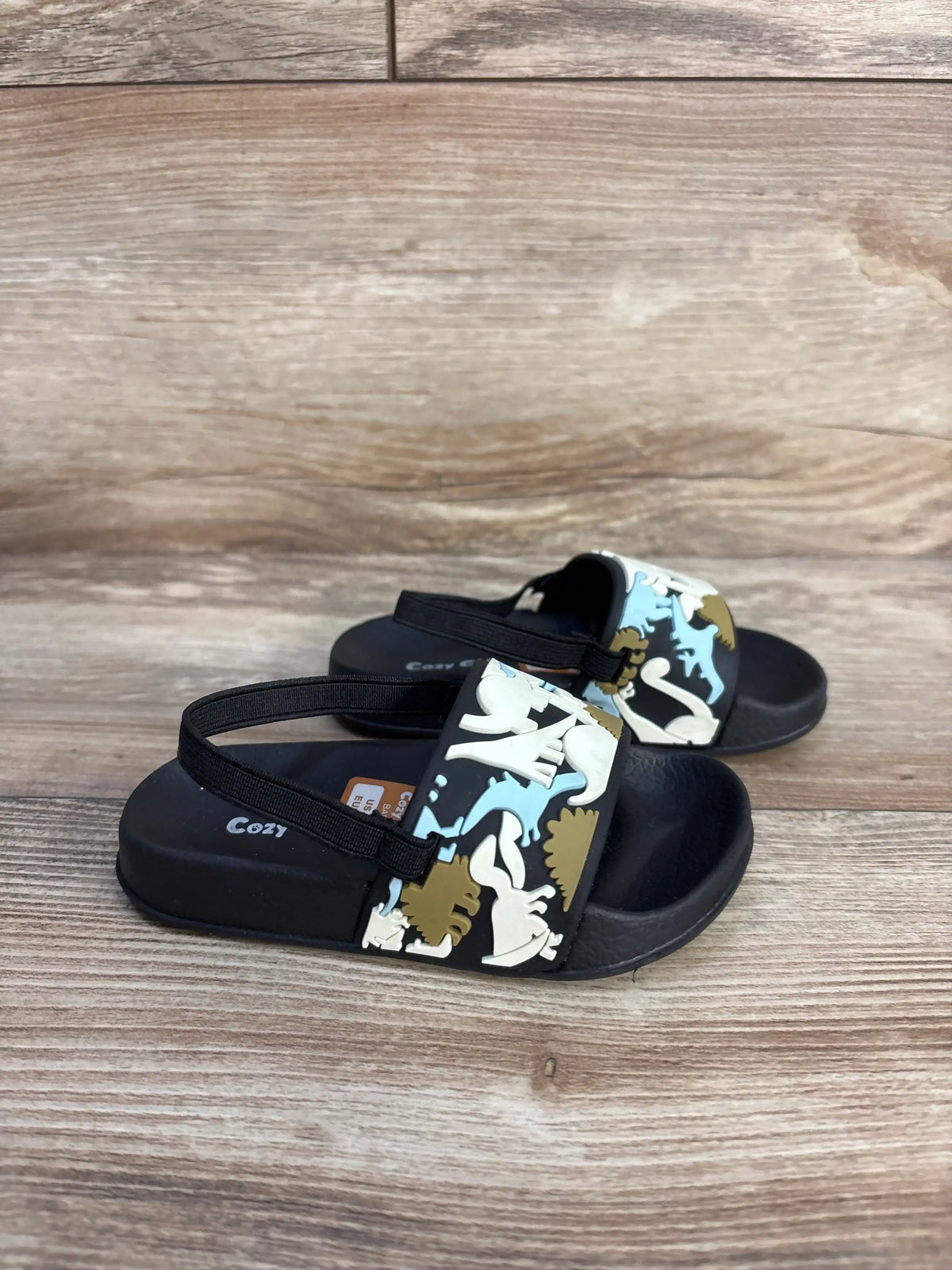 Front three-quarter view of Cozy Cub dinosaur slide sandals showcasing dinosaur-print straps on a wood-grain background