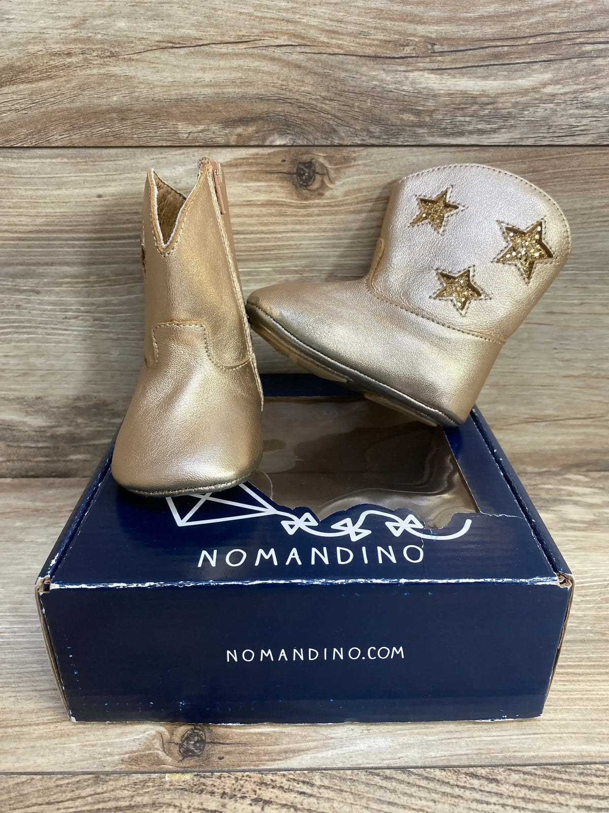 Front and side view of Nomandino infant gold boots showcasing glitter star cutouts and side zipper on navy Nomandino box with wood background