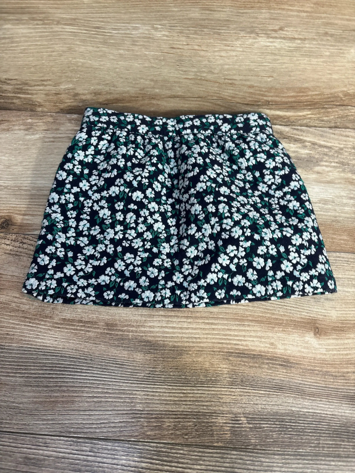 Front flat-lay of toddler black floral skirt with elastic waistband and small white flowers and green leaves on wooden background