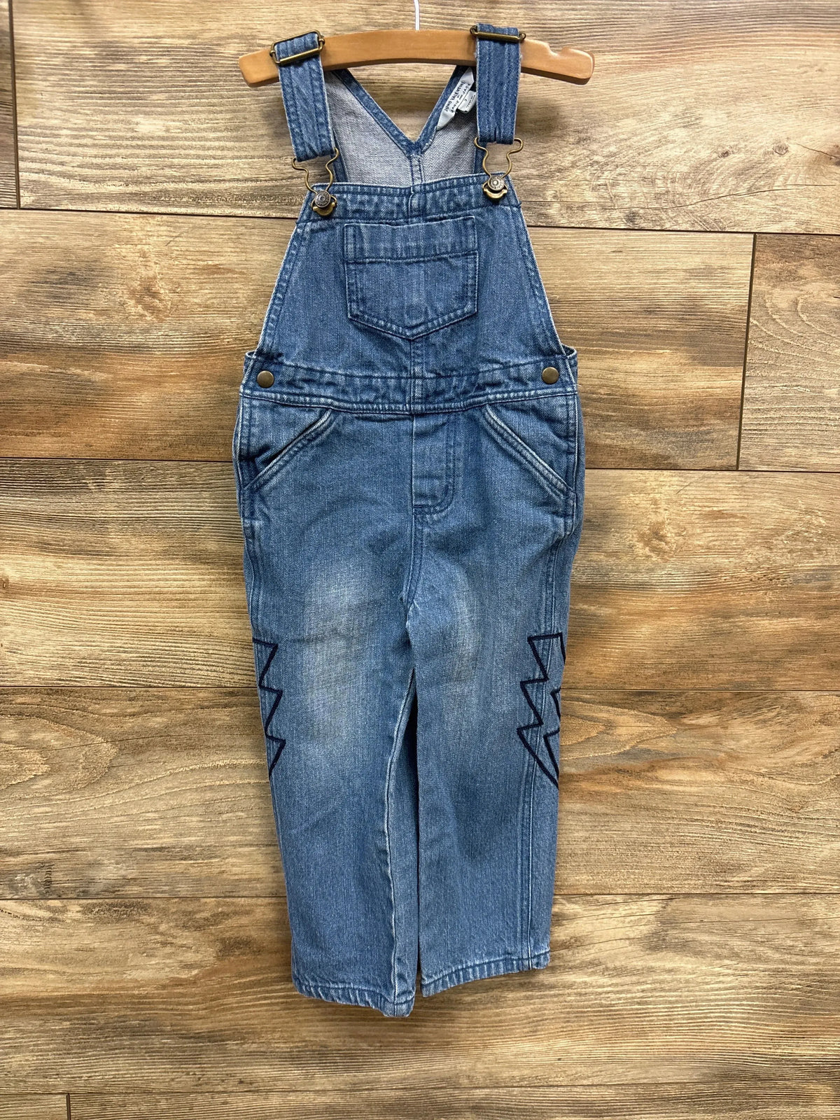 Front of Juno Valentine blue denim overall showing bib pocket, straps and zigzag embroidery at knees on wooden hanger