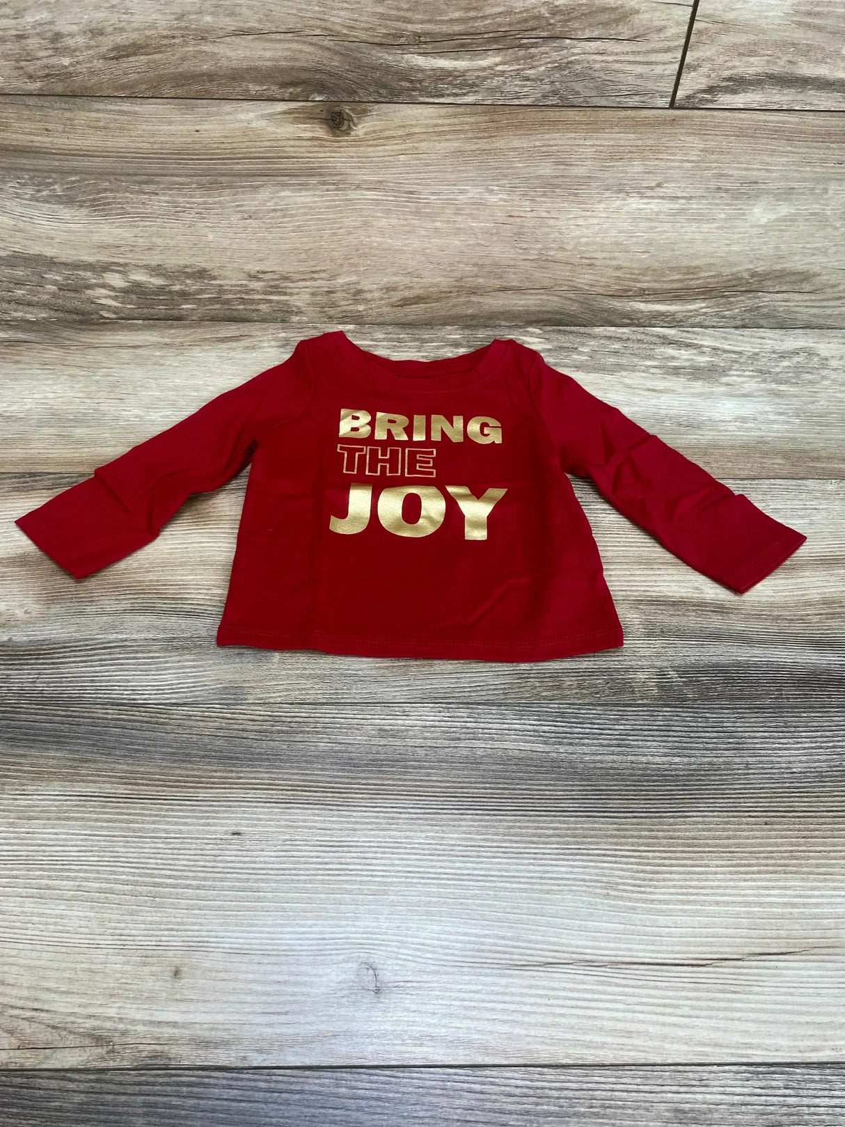 Flat lay front of red Cat and Jack long sleeve baby shirt showing gold BRING THE JOY print on wood floor background