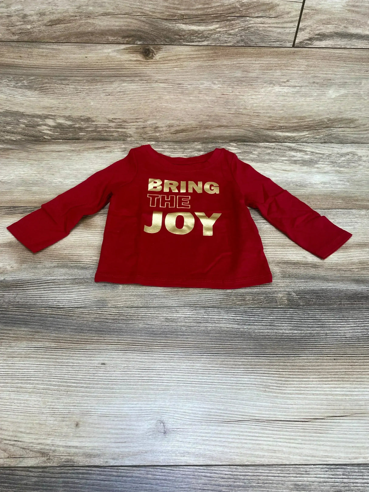 Flat lay front of red Cat and Jack long sleeve baby shirt showing gold BRING THE JOY print on wood floor background