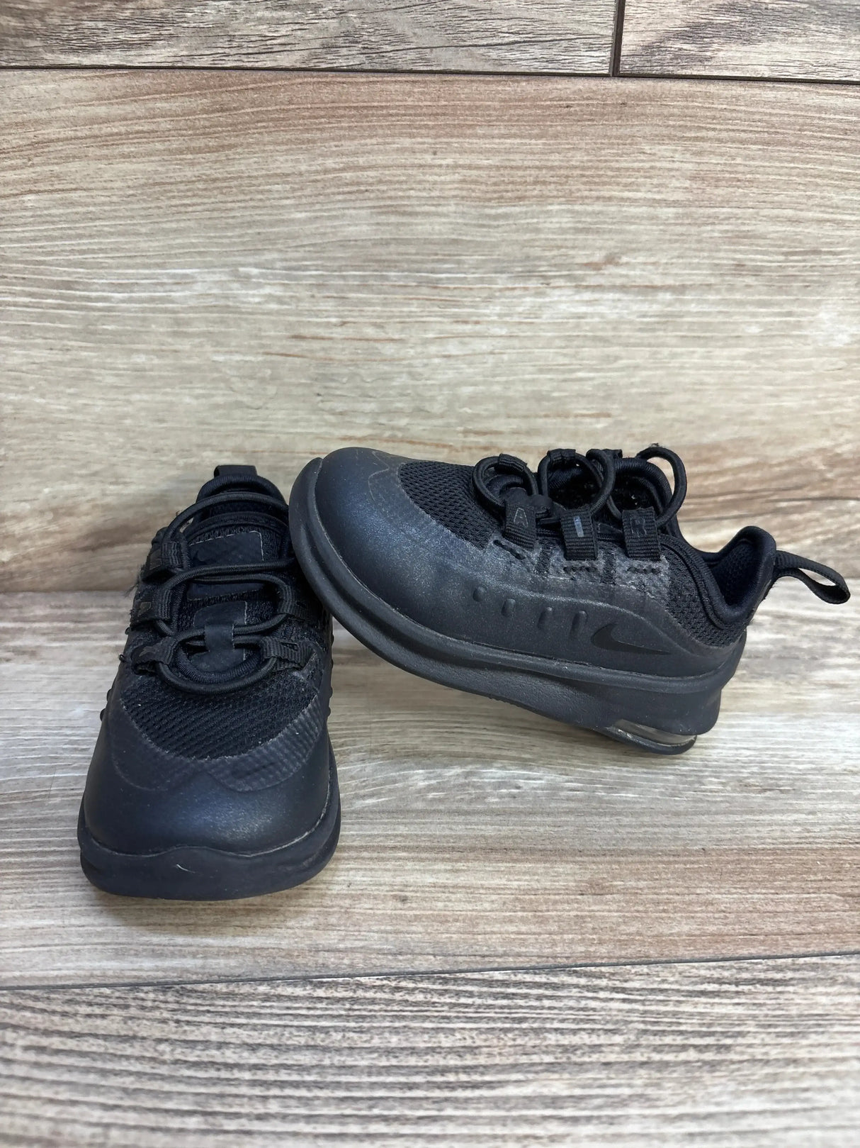 Angled view of toddler Nike Air Max Axis black sneakers with bungee lacing and visible heel air on wood background