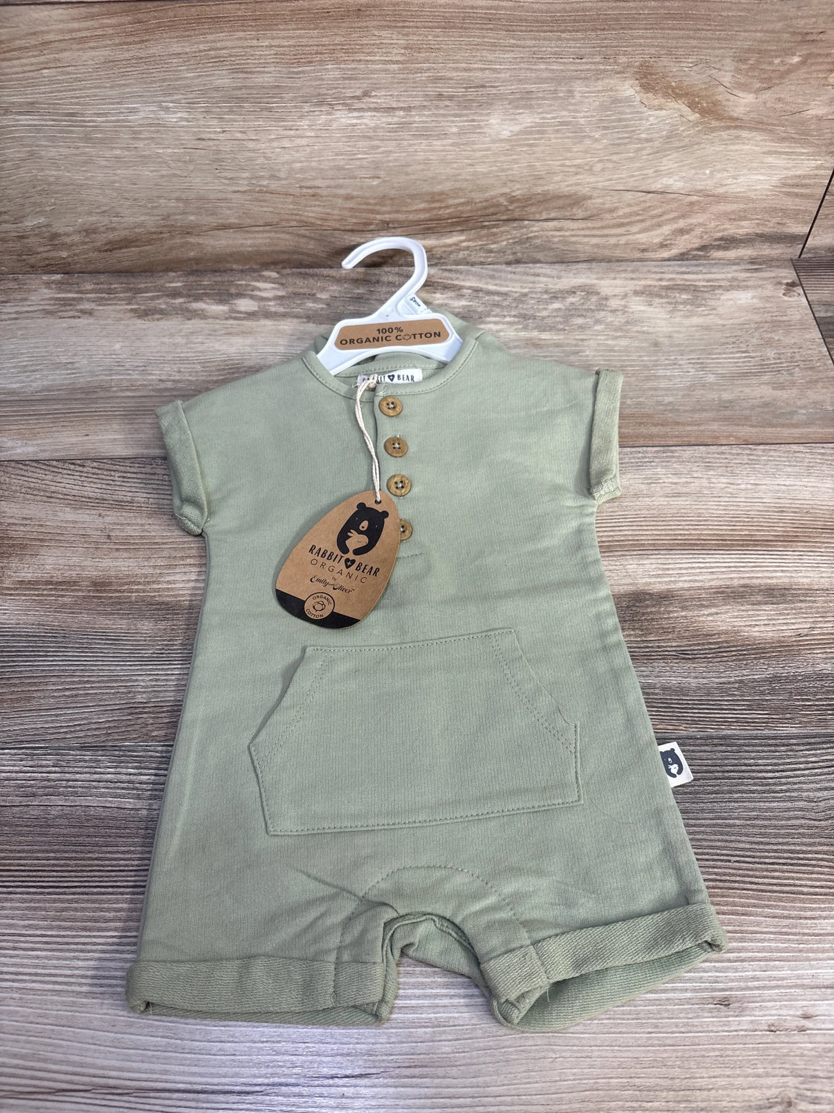 Front view of green Rabbit and Bear organic henley shortie romper showing three wooden buttons, kangaroo pocket, and brand tag on wood background