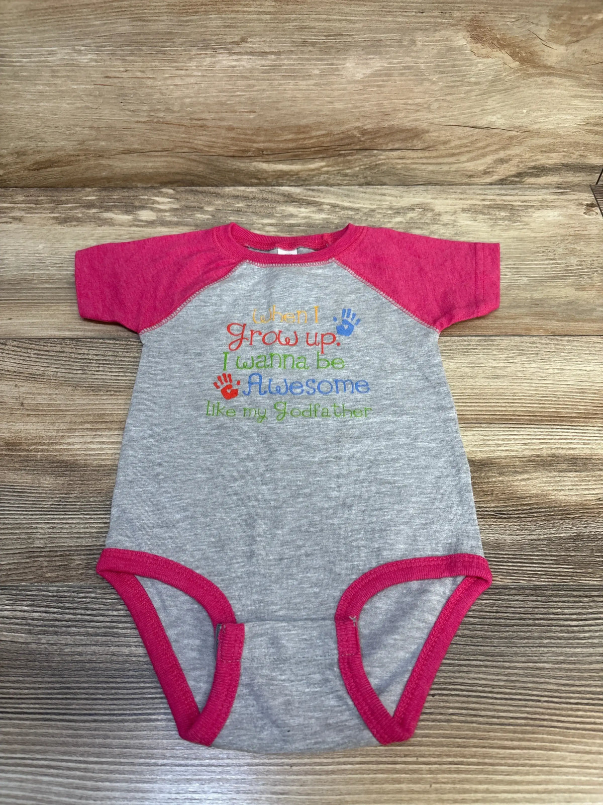 Flat lay of gray baby bodysuit with pink sleeves and trim, embroidered I wanna be awesome like my Godfather on wood background