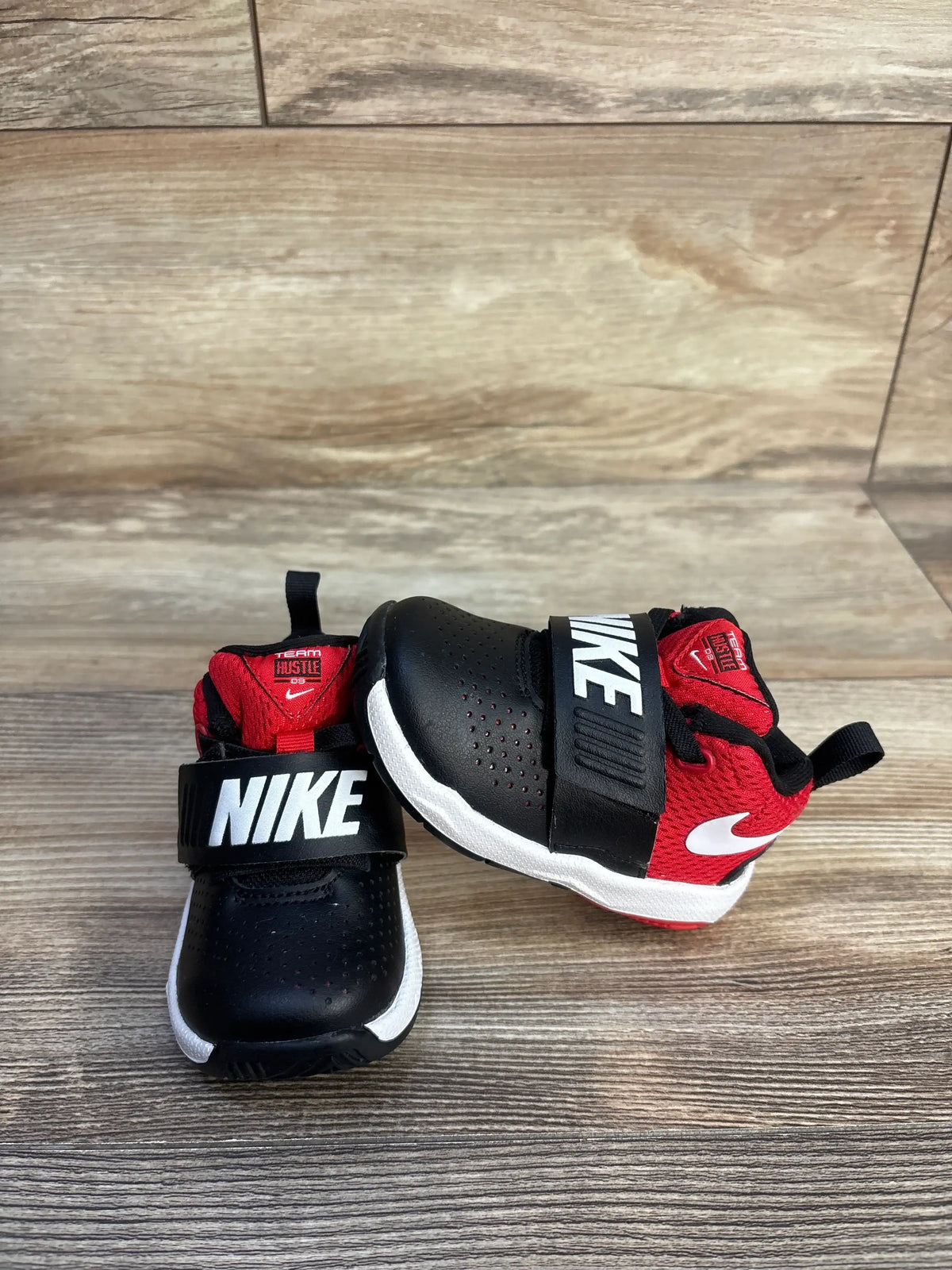 Top and side view of Nike Team Hustle D8 sneakers in black and university red with strap and white midsoles on wood floor
