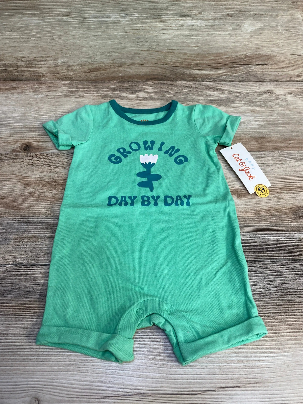 Front view of green Cat and Jack shortie romper showing Growing Day By Day flower graphic and snap crotch on wooden background