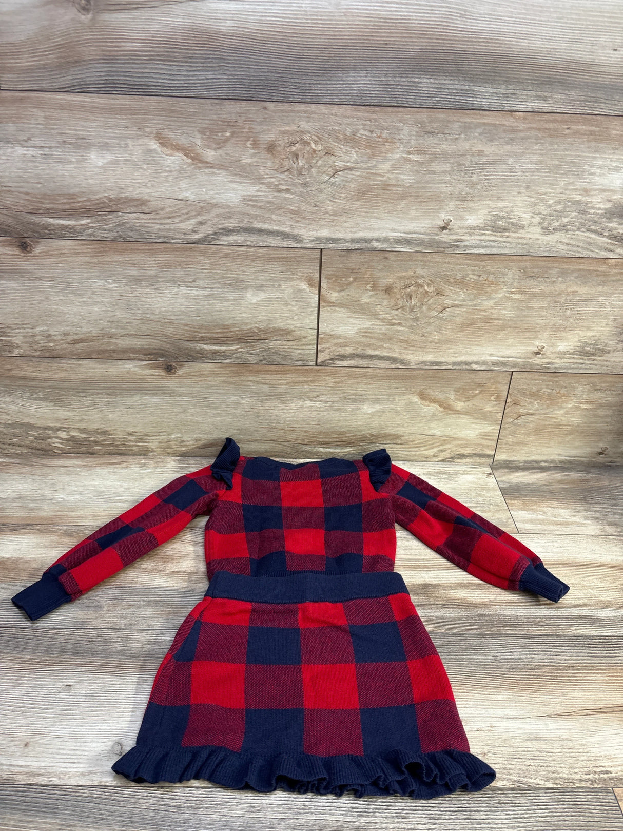 Flat lay of red and navy plaid toddler sweater and matching skirt, showing ruffle shoulders and ruffled hem on wood floor